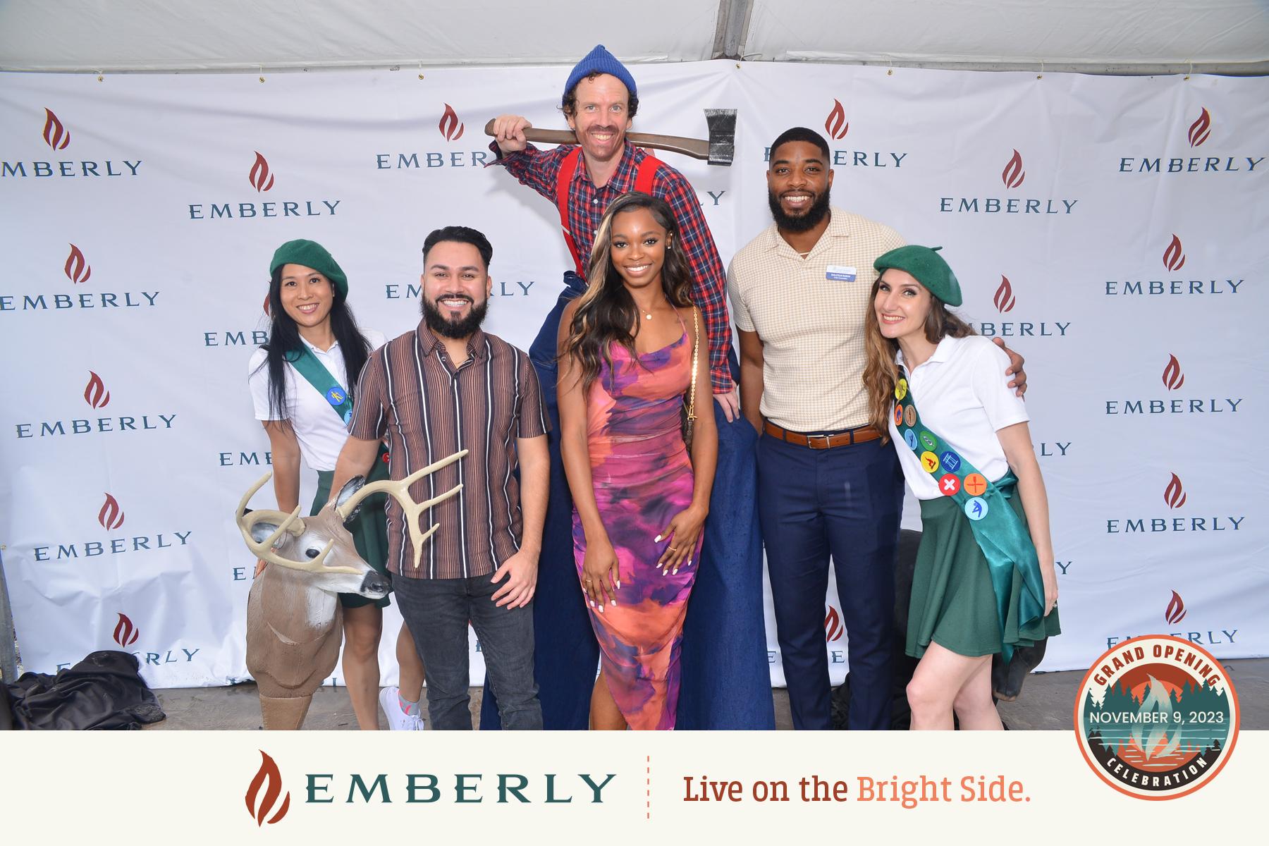 A group of six people posing for a photo in front of an Emberly backdrop. Two are in costumes, one on stilts and another with a deer head mask. Text reads: "Emberly - Live on the Bright Side." Discover new homes in our master planned community!