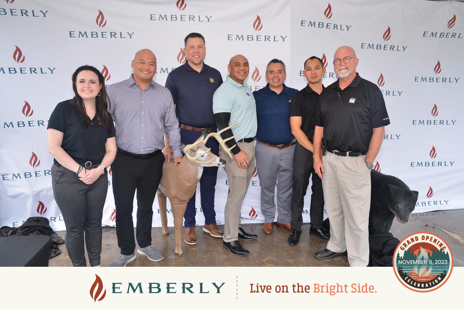 A group of seven people, including one holding an archery deer target, are standing and smiling in front of an "Emberly" banner. A "Grand Opening" emblem with the date November 9, 2023, is displayed, celebrating new homes in Fort Bend County.