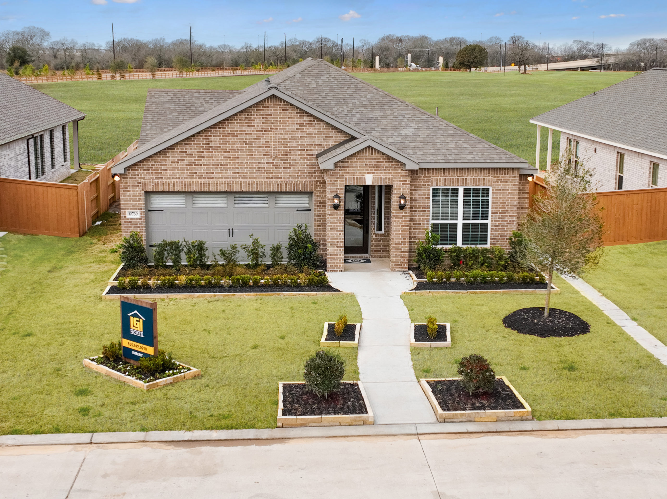 Single-story brick house with a two-car garage and manicured front yard, including shrubs and plants along the walkway. Located near Rosenberg in Fort Bend County, an Emberly for-sale sign is placed on the left side of the lawn.