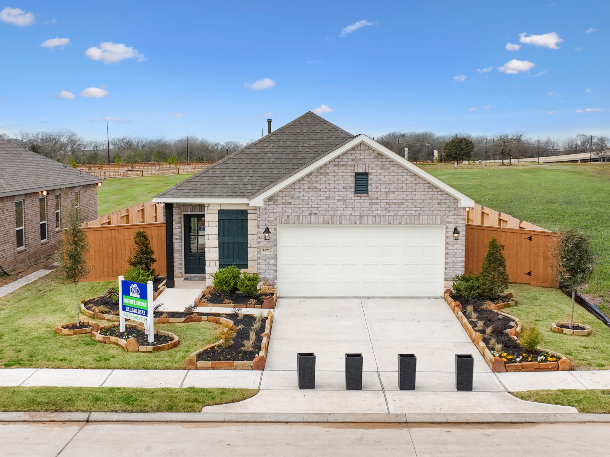 A single-story brick house with a two-car garage and well-maintained front yard near Rosenberg. A "Model Home" sign is displayed in the yard. The house, located in Emberly, is surrounded by a wooden fence and sits on a quiet street in Fort Bend County.