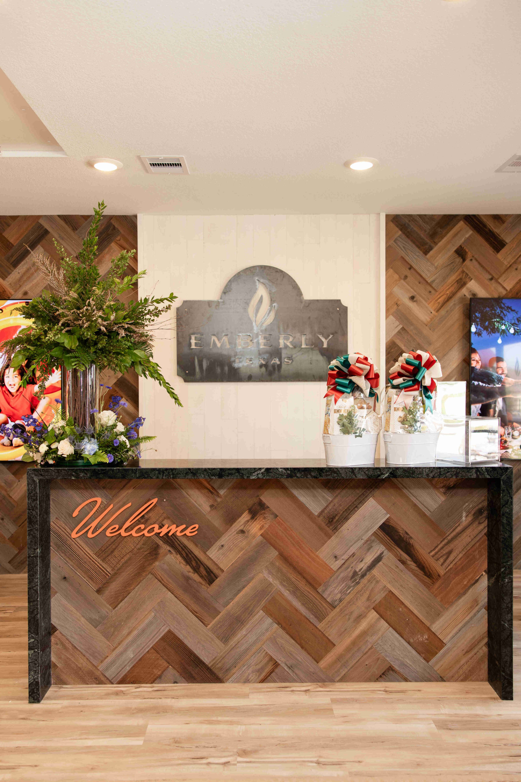 Reception desk with a "Welcome" sign, decorated with plants and floral arrangements, in front of a wall-mounted metallic sign reading "Emerly." Perfect for an event or grand opening, the background features photos and wooden herringbone-patterned panels.