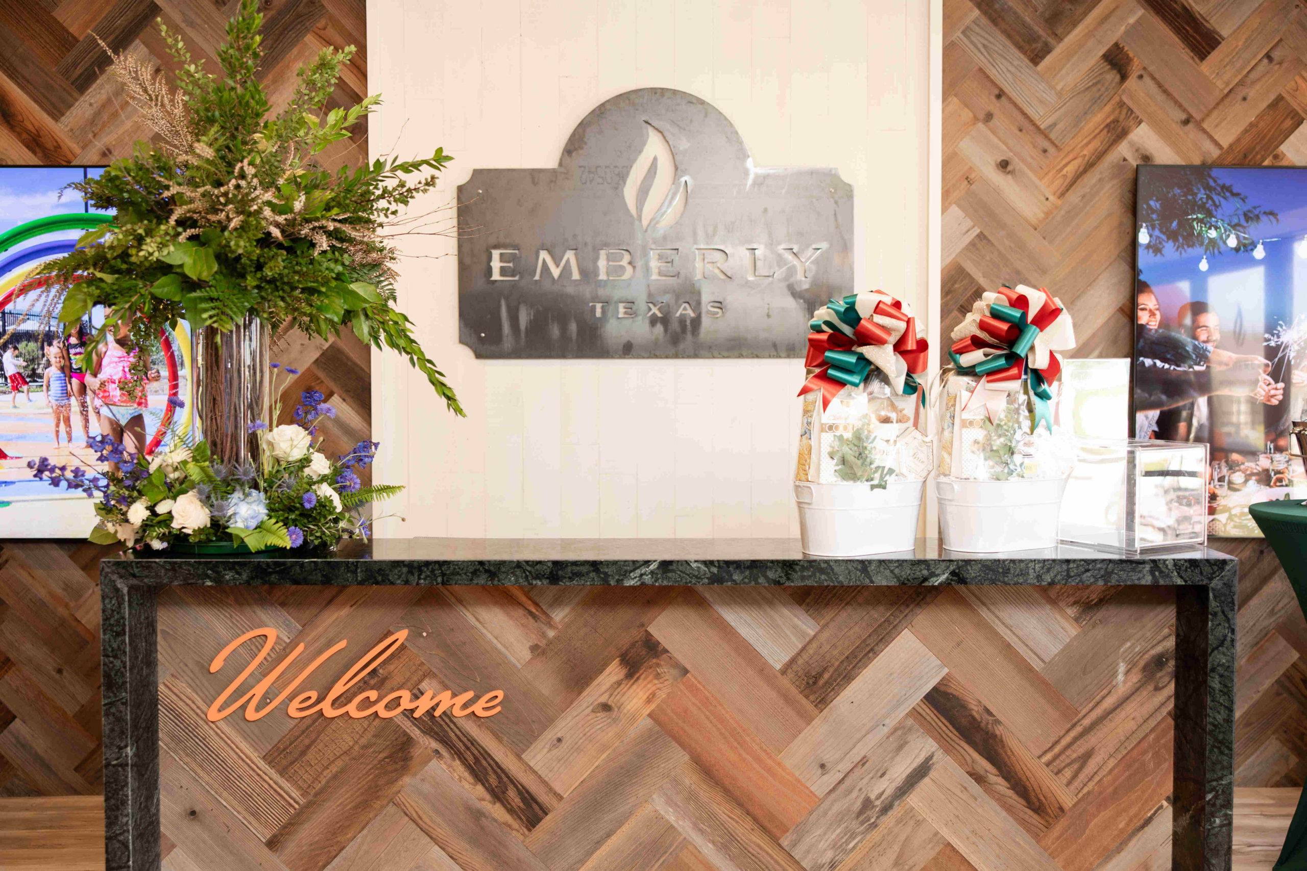 A reception desk with a "Welcome" sign and floral arrangements, featuring a backdrop with the logo "Emberly Texas," sets the stage for our Grand Opening Event.