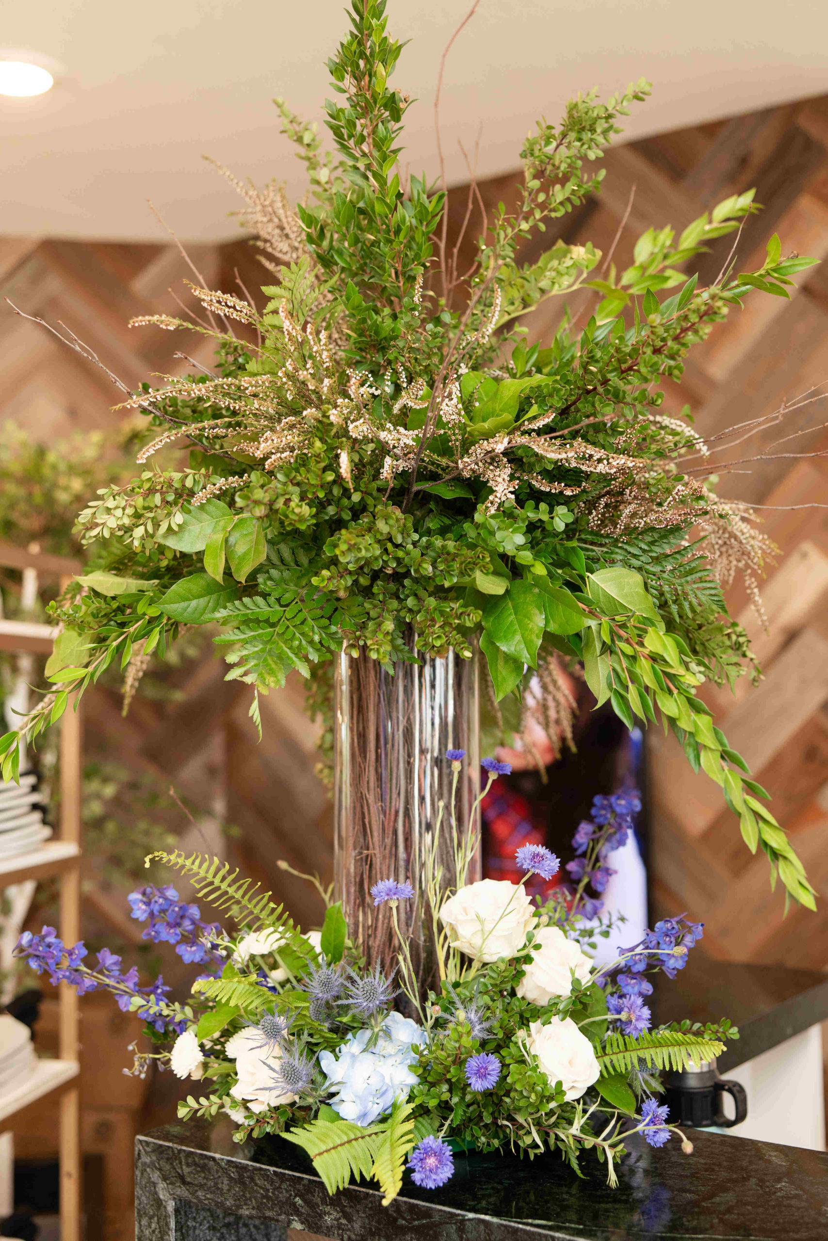 The grand opening floral arrangement features green foliage, white roses, blue flowers, and brown branches in a tall, clear vase on a wooden-patterned background.