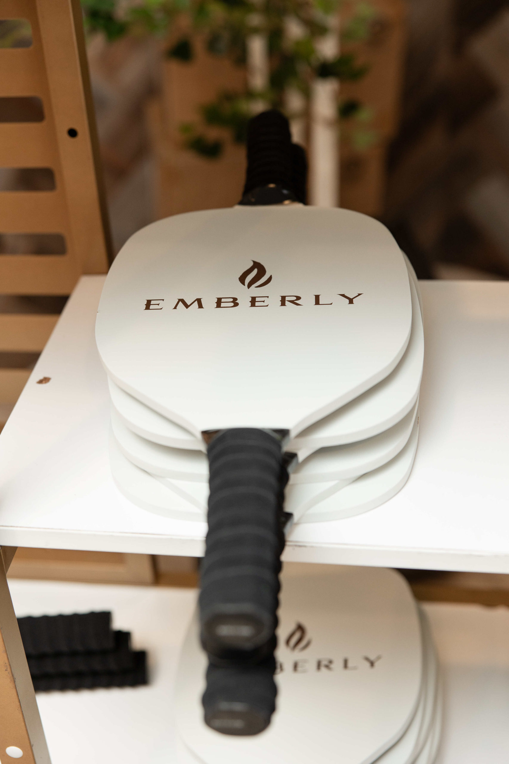 A stack of white pickleball paddles branded "Emberly" with black grips rests on a white shelf, ready for the Grand Opening Event at the Information Center.