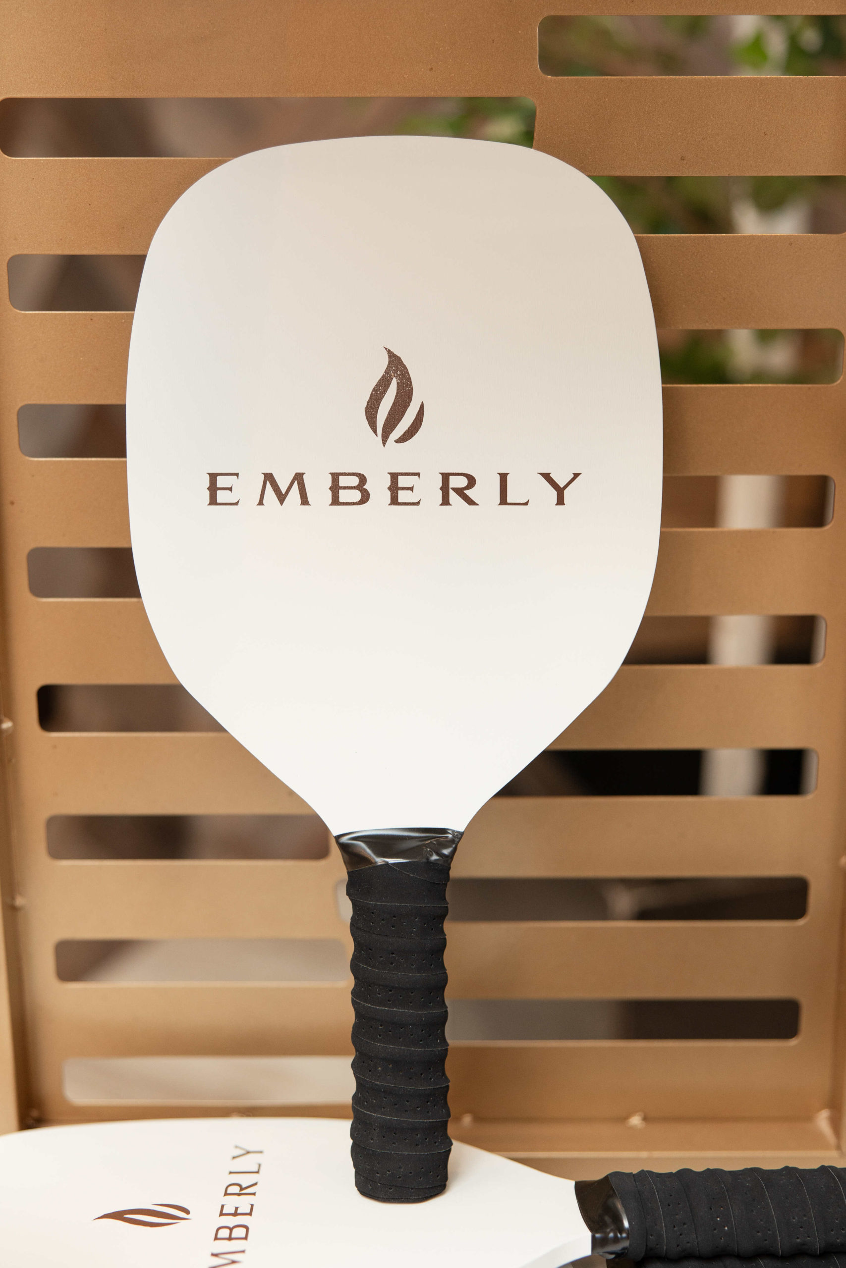 A pickleball paddle resting against a wooden slat, featuring the brand name "Emberly" and a flame logo on the face. The paddle has a black grip and a white surface, perfect for showcasing at the Information Center Grand Opening event.