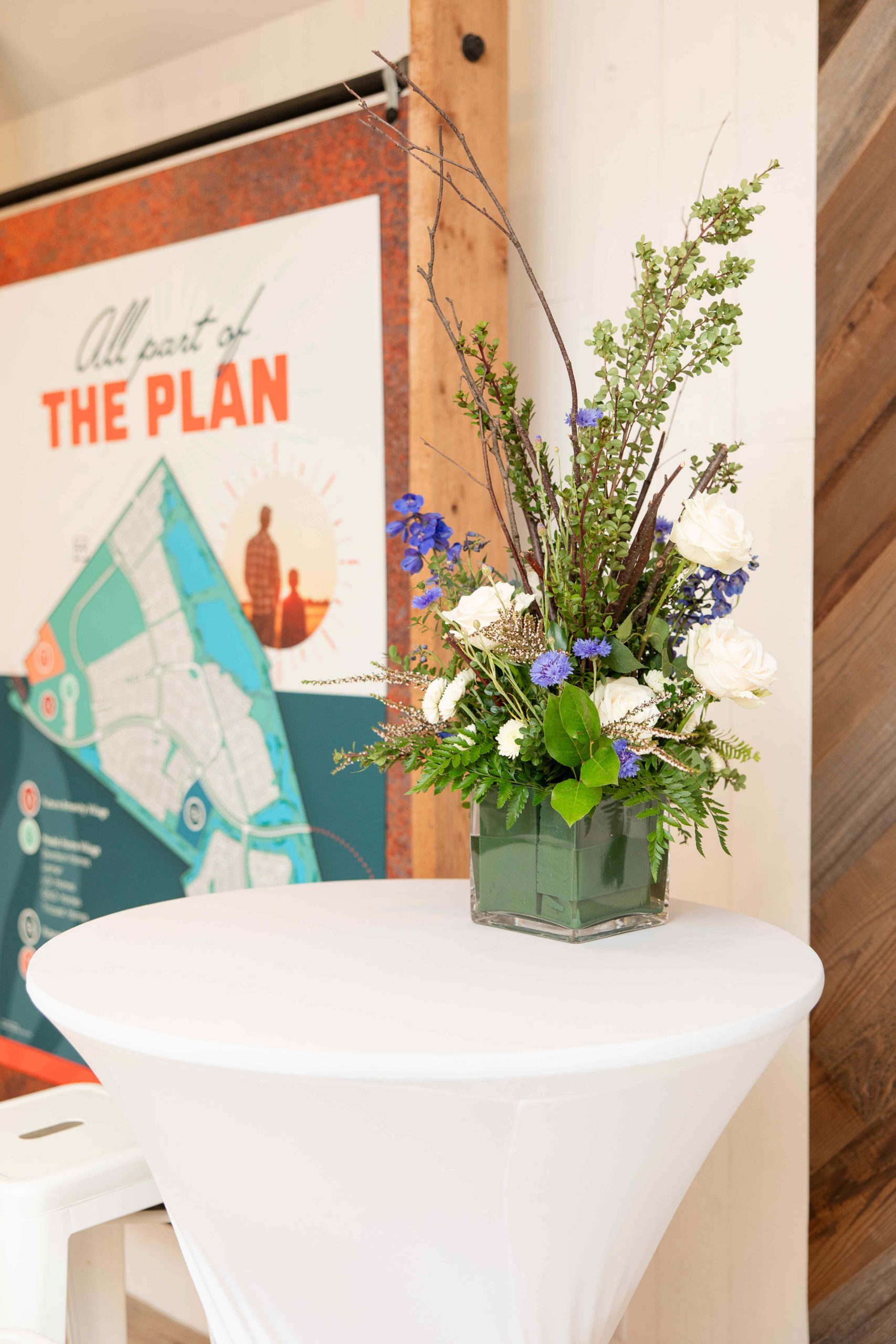 A small floral arrangement in a square vase sits on a white-covered cocktail table in front of a poster featuring a map and the text "All part of THE PLAN" at the grand opening.