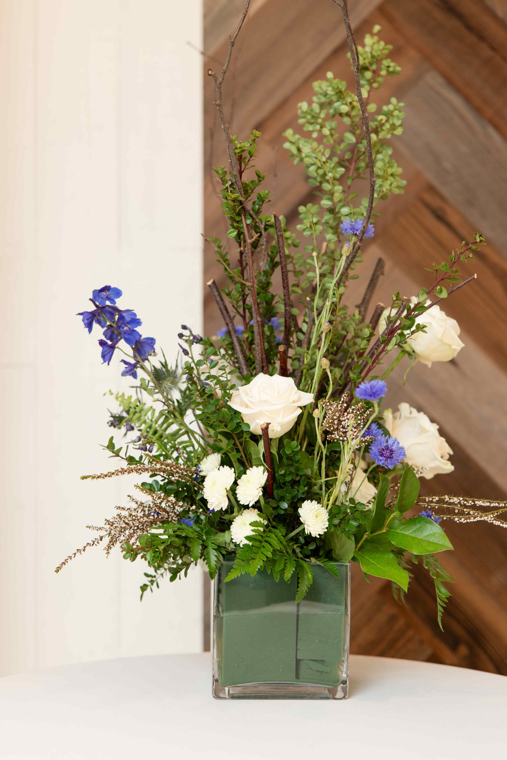 A floral arrangement with white roses, purple flowers, white chrysanthemums, green leaves, and twigs in a square glass vase sits elegantly on a table. A wooden herringbone patterned wall forms the backdrop. Perfect for an event or grand opening at an information center.