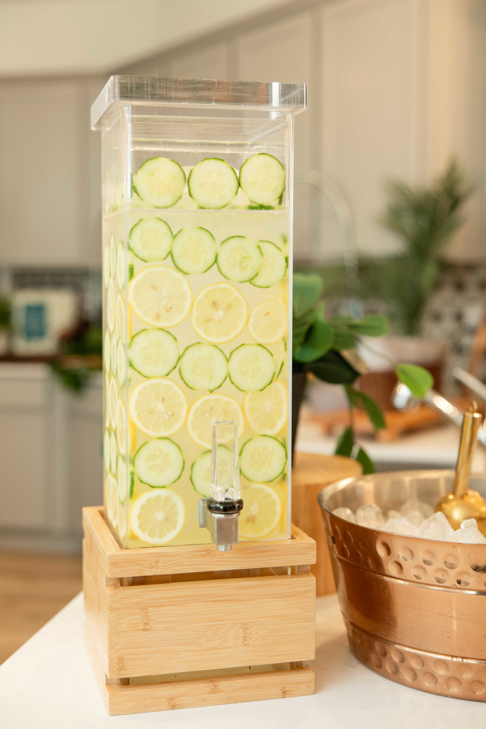 A clear beverage dispenser filled with water infused with cucumber and lemon slices, placed on a wooden stand next to a copper ice bucket, graced the kitchen counter for the Grand Opening Event.