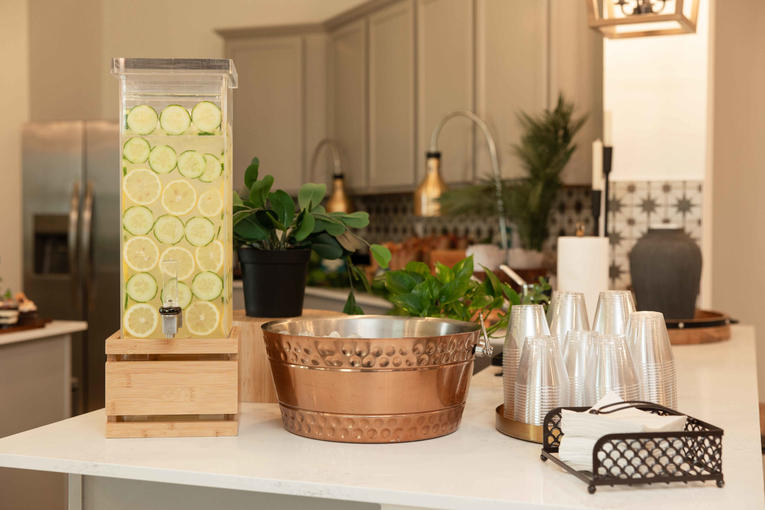 A kitchen countertop displaying a beverage dispenser filled with water and lemon slices, a copper ice bucket, stacked plastic cups, and a napkin holder for the grand opening event, with a plant and kitchenware in the background.