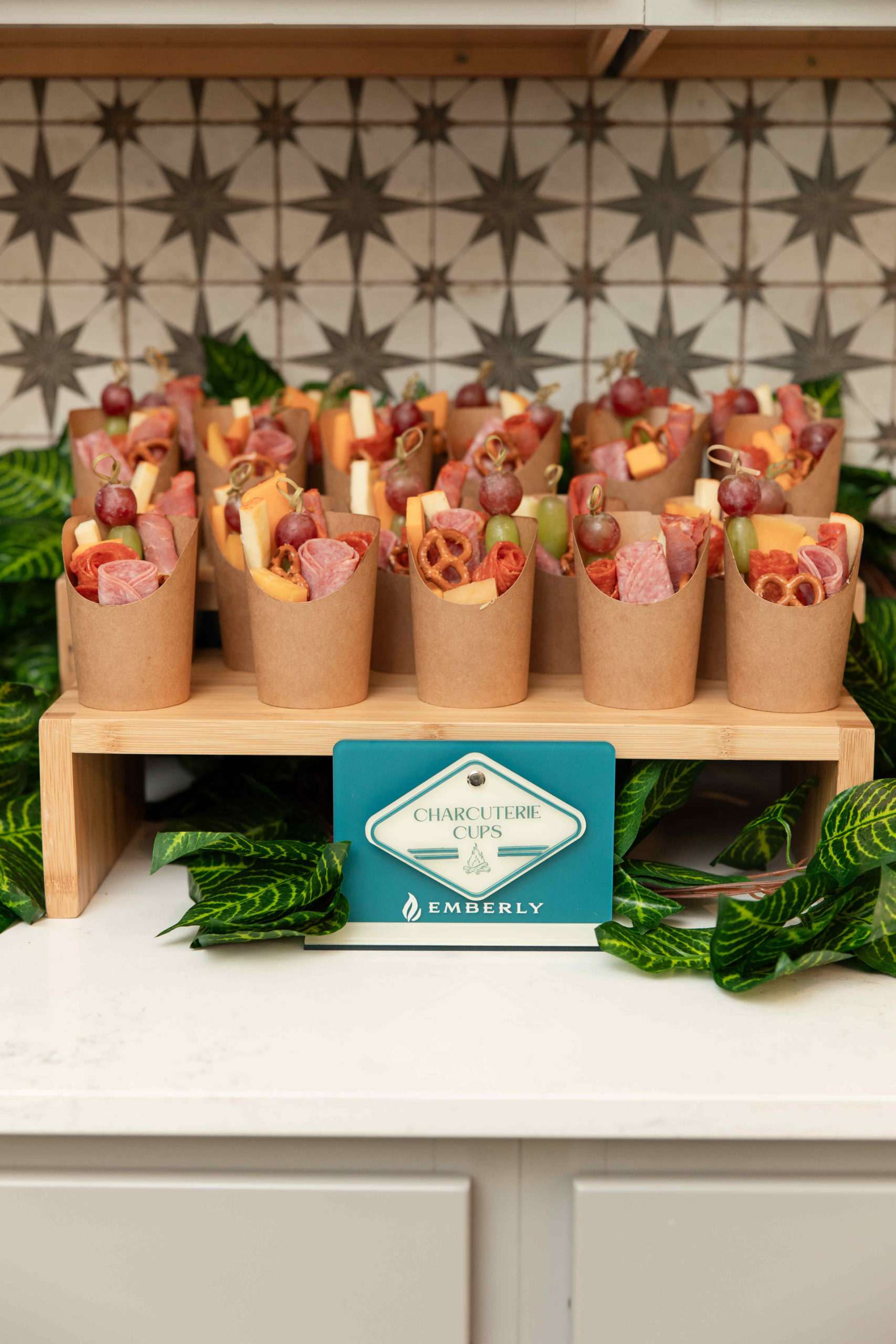 A display of charcuterie cups on a wooden stand with a sign reading "Charcuterie Cups by Emberly" is surrounded by green leaves, elegantly designed for the grand opening event. The cups contain various meats, cheeses, and snacks.