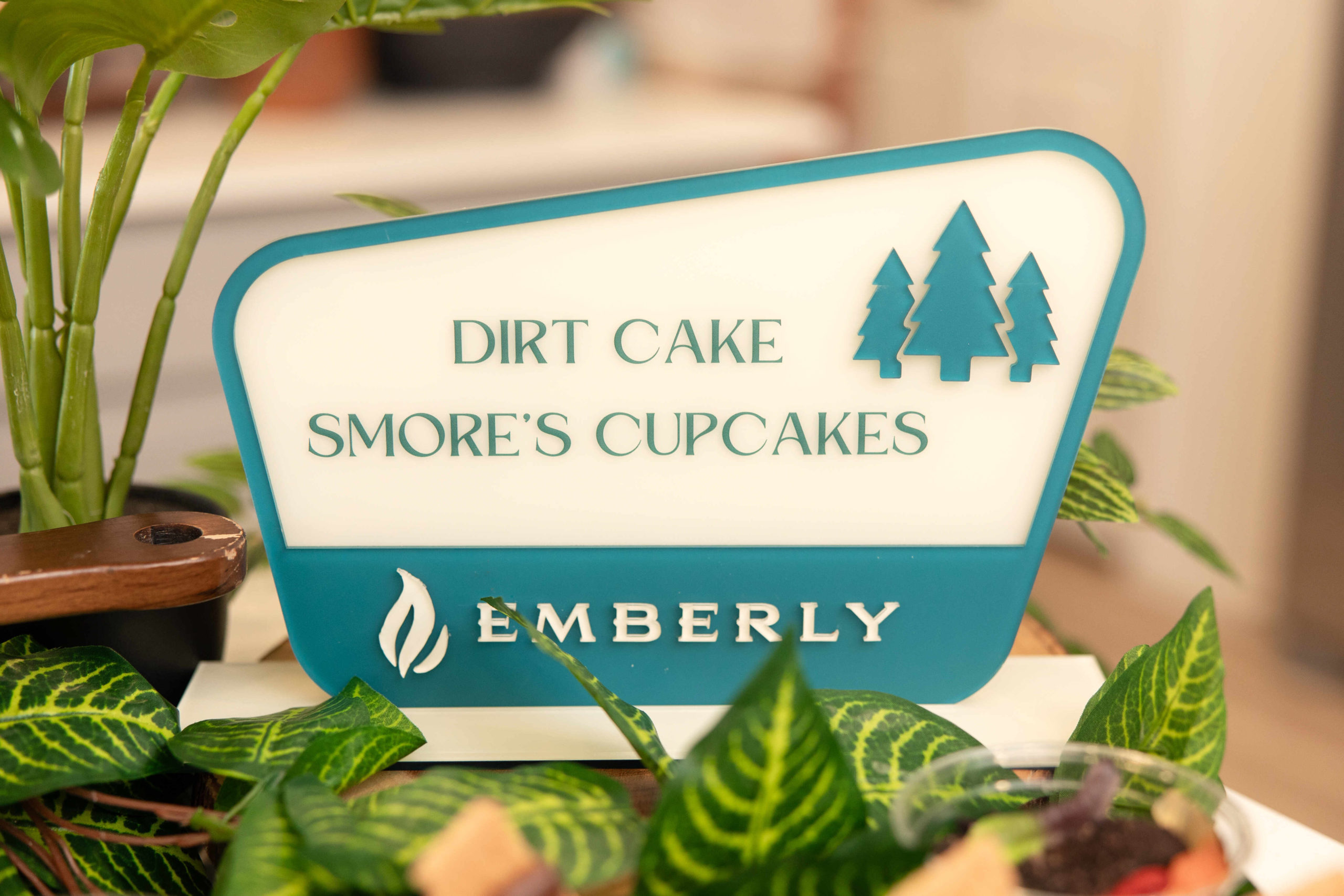 A sign that reads "Dirt Cake S’mores Cupcakes Emberly" with illustrations of trees and a flame, surrounded by plants, marks the grand opening event at our Information Center.