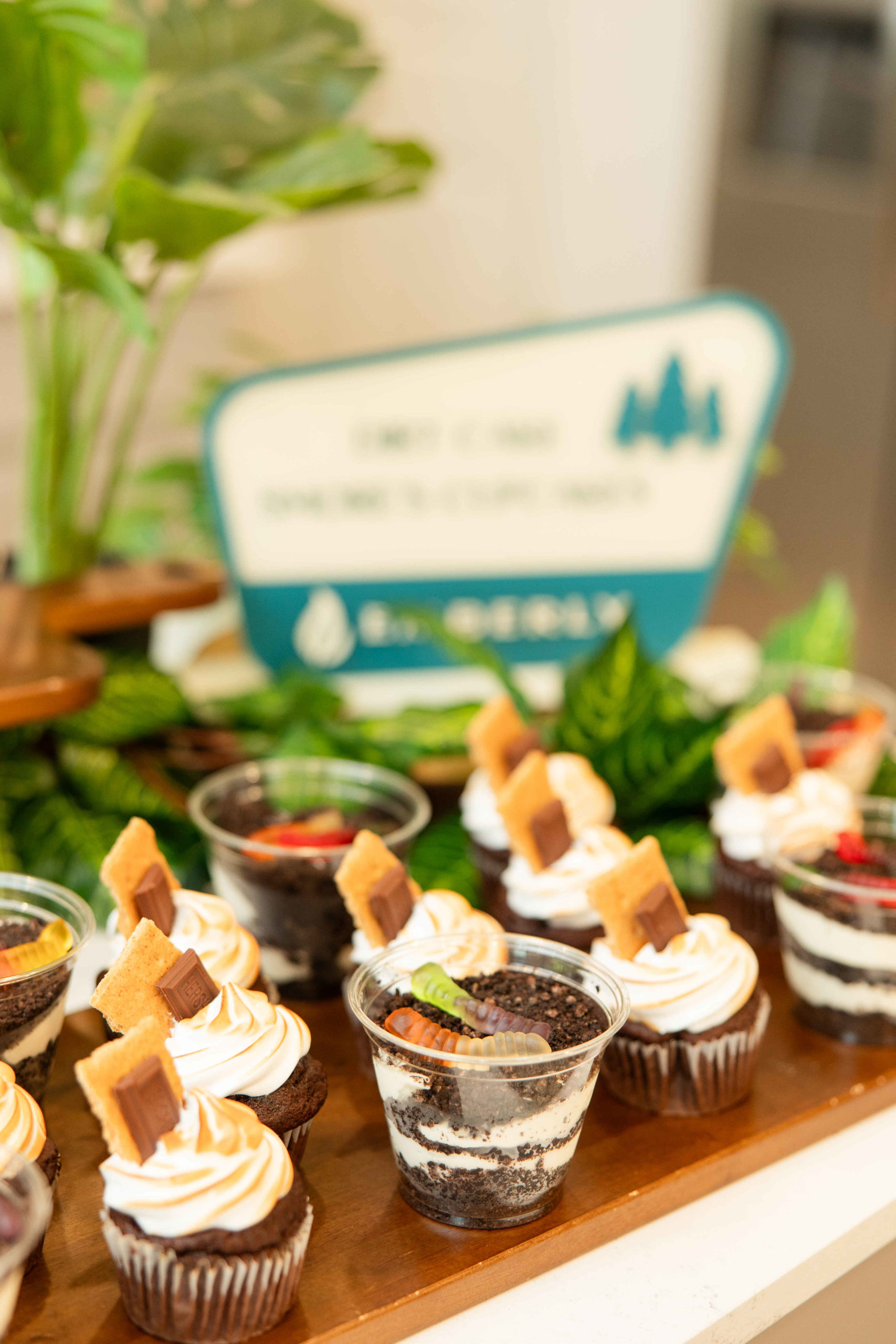 Close-up of several dessert cups displayed on a tray at the Grand Opening event. The dessert cups have layers of chocolate and cream, topped with cookies and whipped cream. A blurred sign and plants in the background hint at the nearby Information Center.