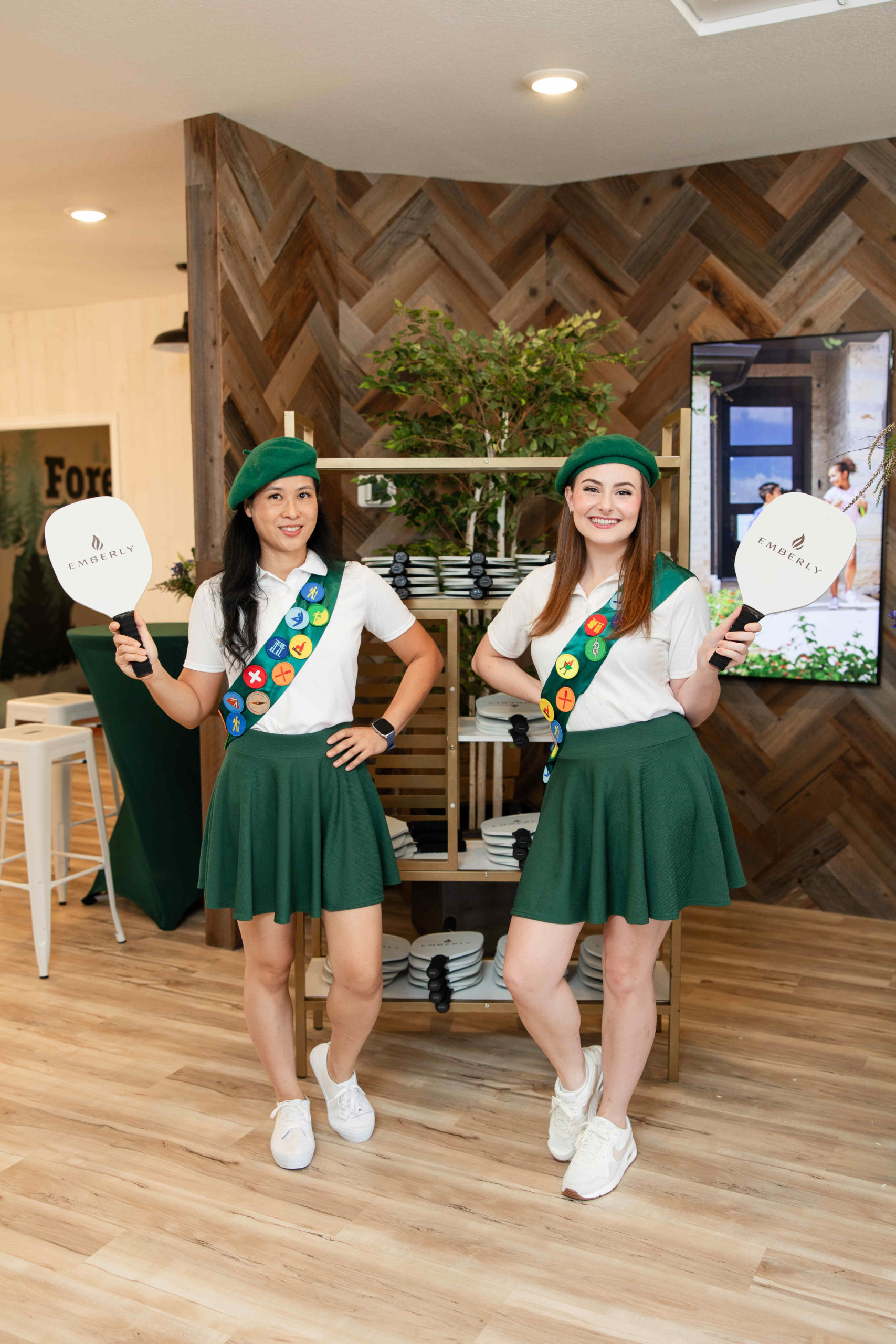 Two people in green skirts, white shirts, and green hats, wearing sashes with colorful badges, hold handheld signs that read "EMERALD CITY" as they pose inside a room with wooden walls at the grand opening event of the Information Center.