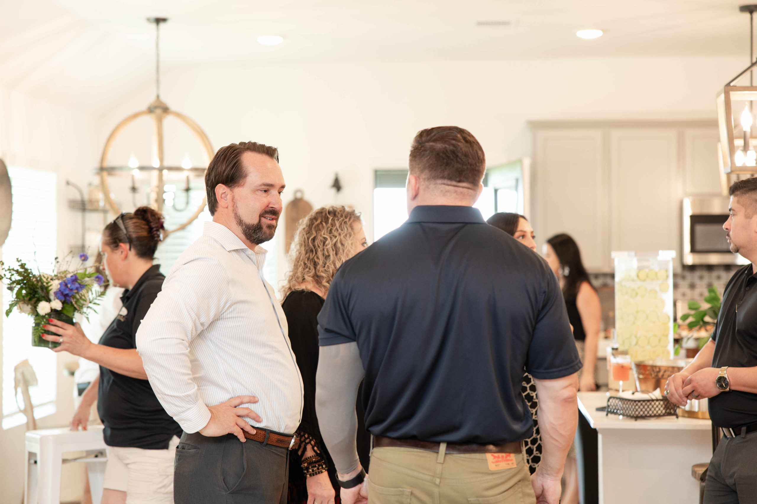 A group of people stand and converse in a well-lit kitchen at the grand opening. One person holds a bouquet, and another is near a table with a lemonade dispenser.