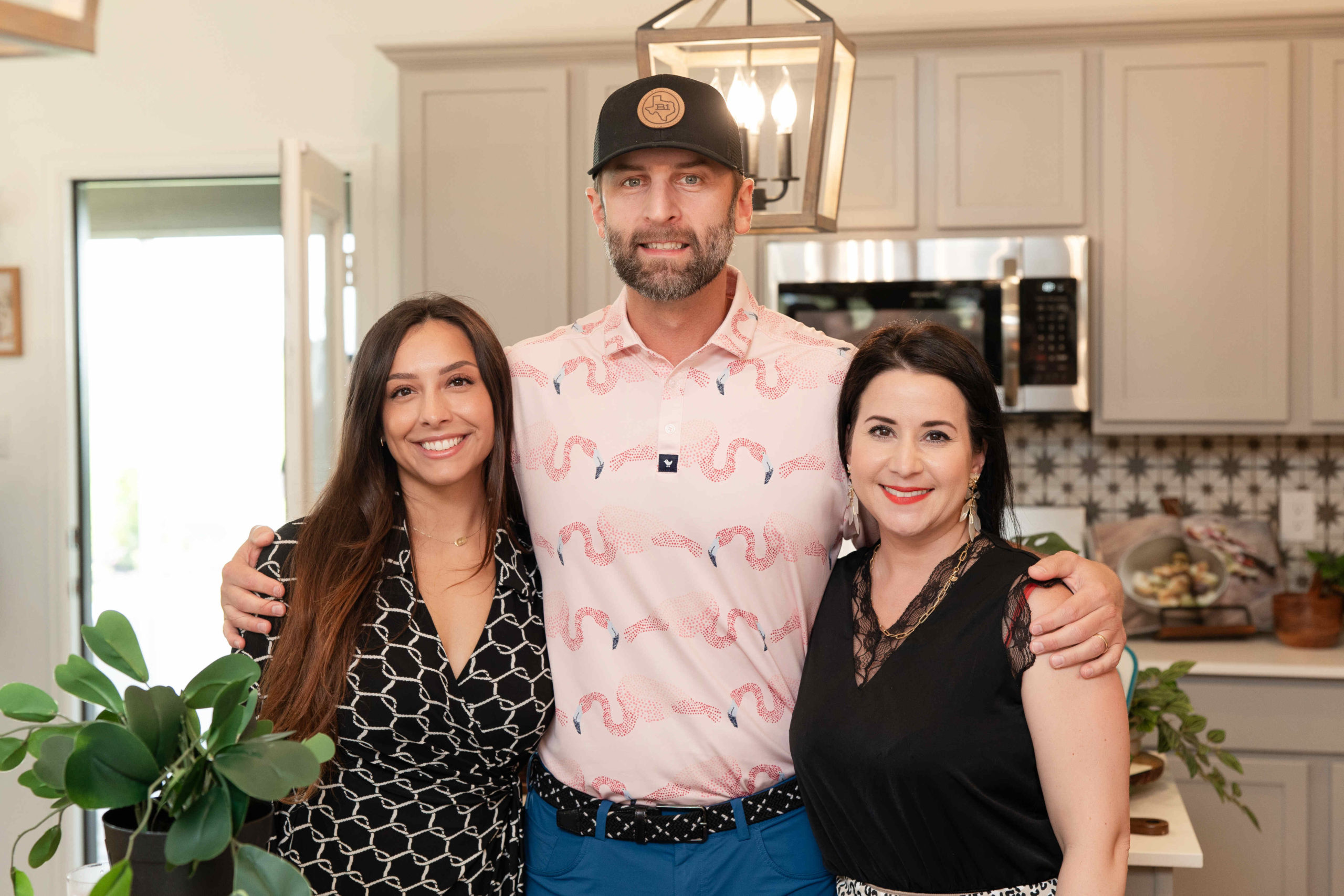 Three people standing together in a kitchen at the grand opening, with the middle person wearing a pink shirt and a black cap, flanked by two smiling women.