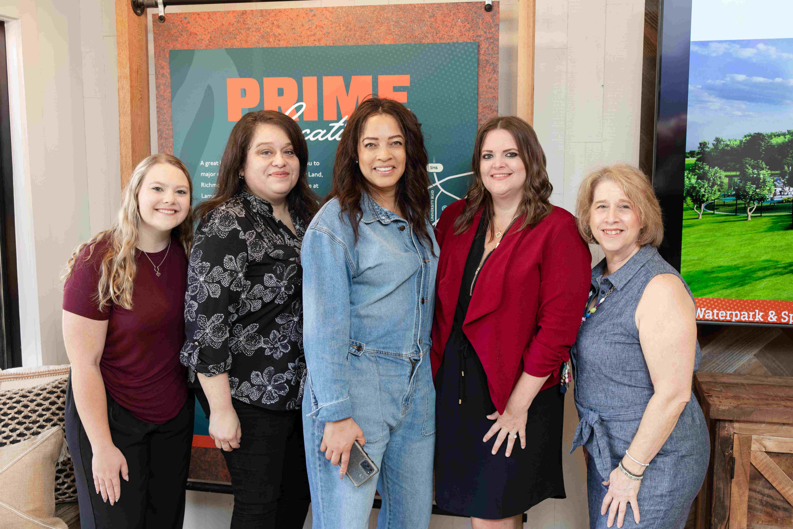 Five women are standing and smiling in front of a sign that reads "PRIME" and a large television displaying a nature scene, celebrating the Grand Opening of the new Information Center.