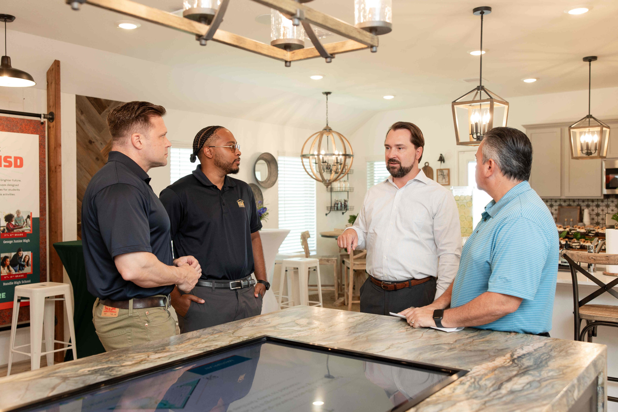 Four men stand in a modern kitchen engaged in conversation during the Grand Opening Event, with one man gesturing while speaking. The kitchen features hanging lights, bar stools, and a stone countertop.