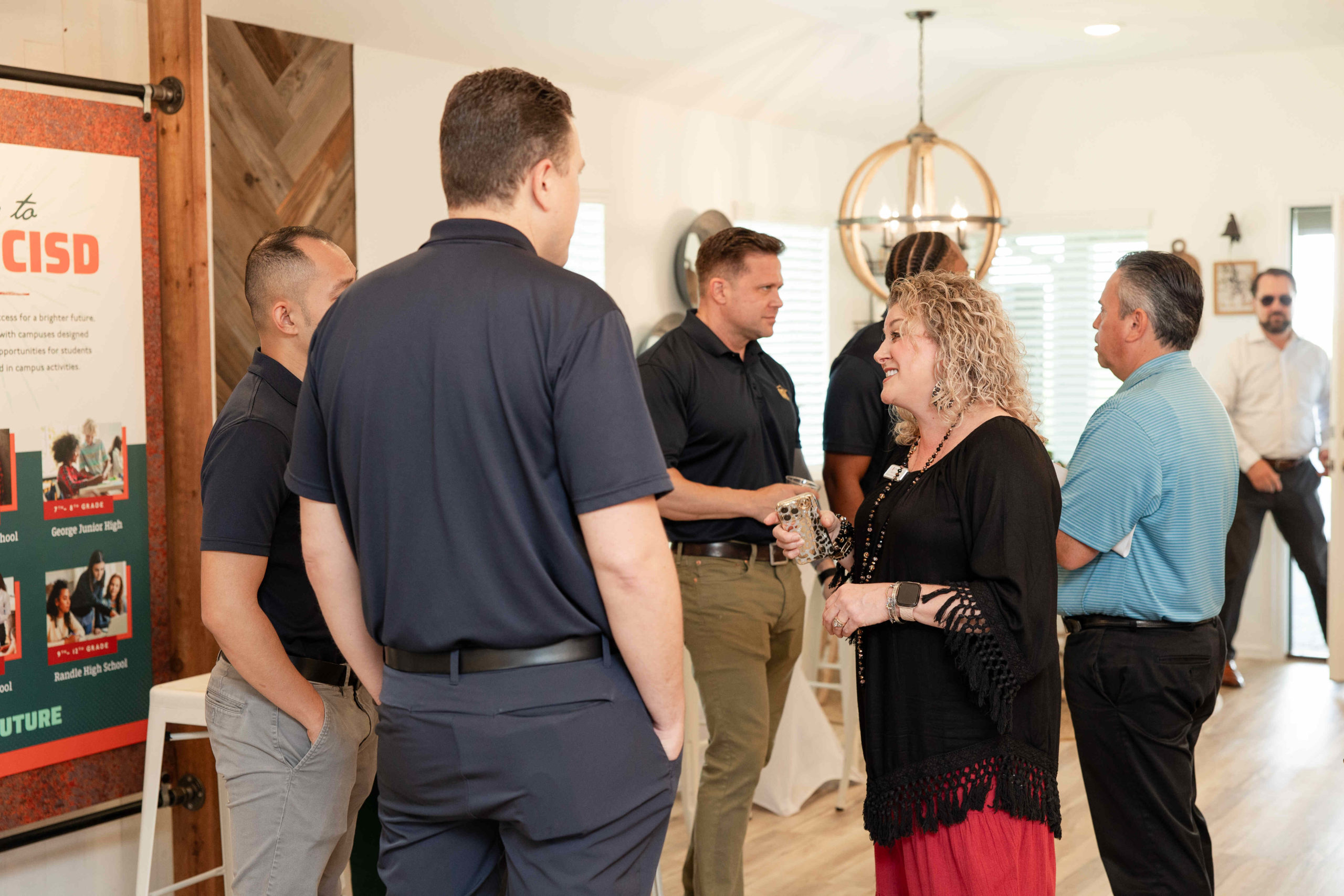 A group of people having a conversation indoors at an information center. One woman and several men are engaged in discussion near a poster. The setting appears informal, as if they are preparing for an upcoming event or grand opening.