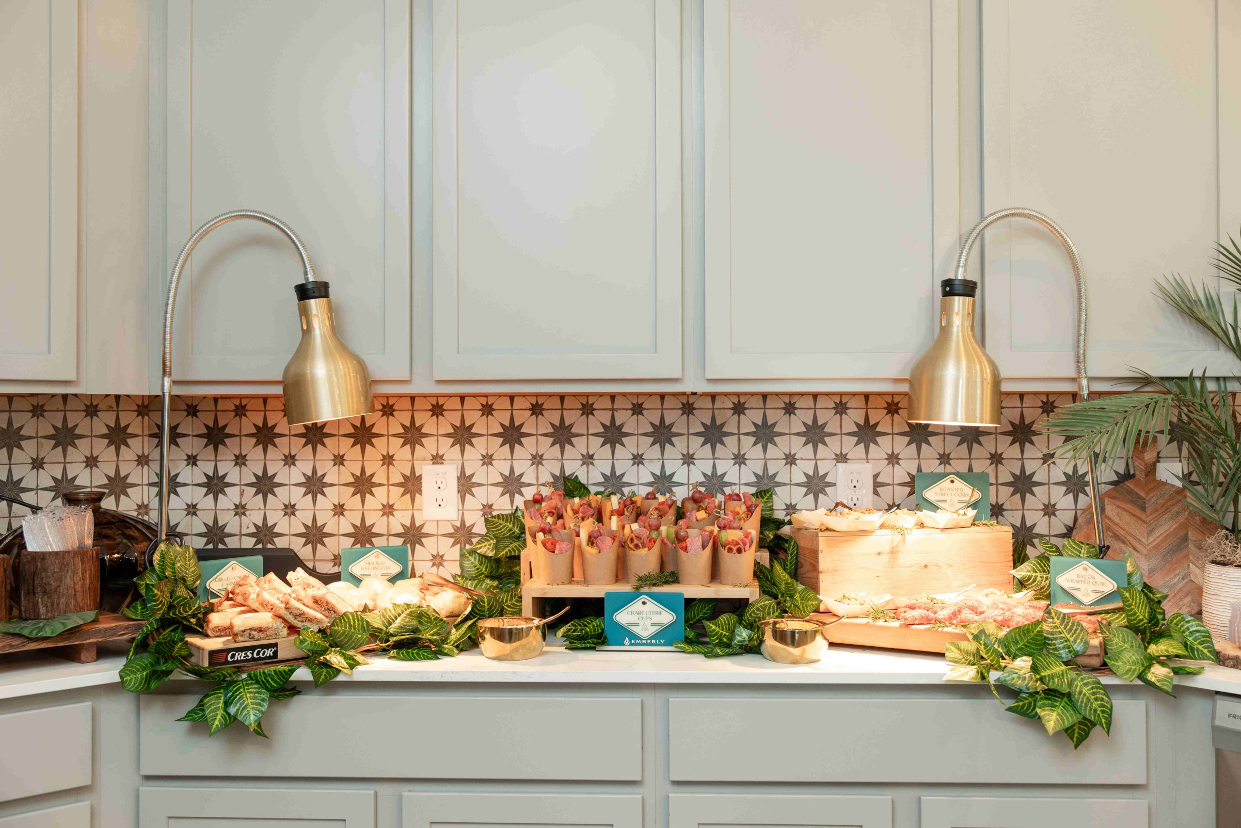 A kitchen counter with a patterned backsplash features food items, including sandwiches and wrapped snacks, under two brass lamps. Green leaves are used as decor around the food, setting the perfect scene for the grand opening event.