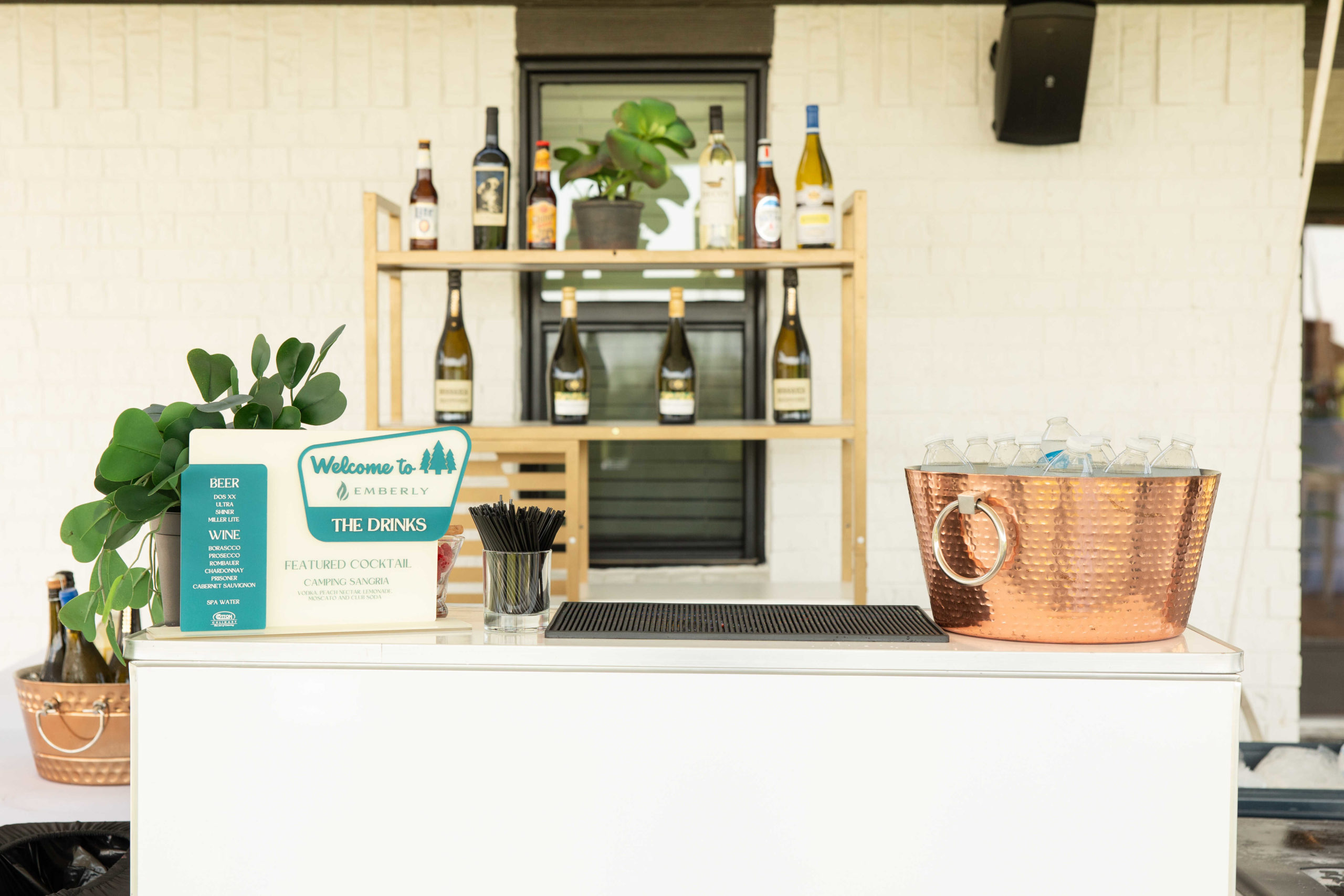 A small bar setup with a shelf of wine bottles behind a white counter, ready for the grand opening. The counter features a sign, plant, straws, and a copper ice bucket filled with bottled drinks.