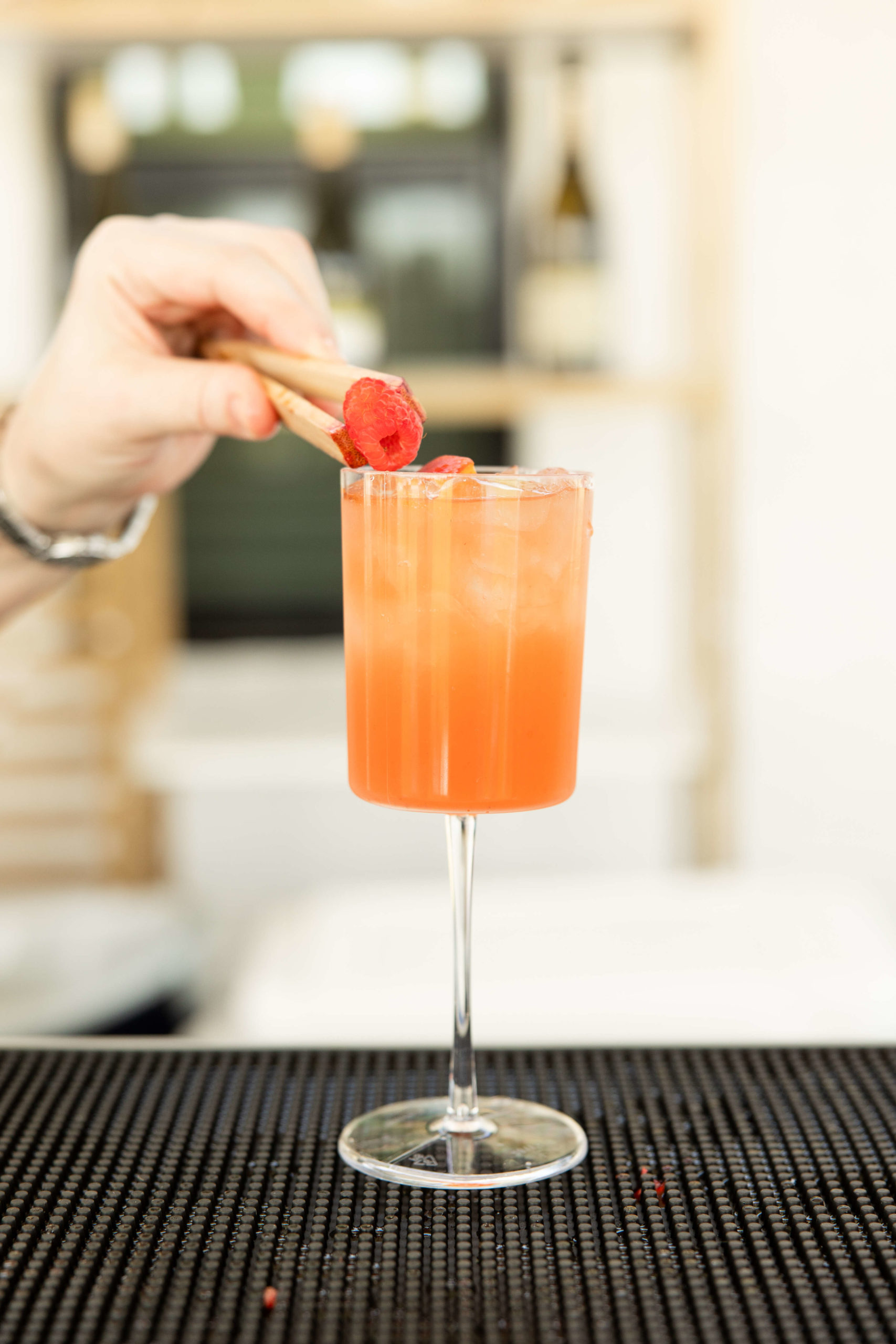 At the grand opening, a hand uses chopsticks to place a garnish on a tall glass of orange-colored cocktail with ice.