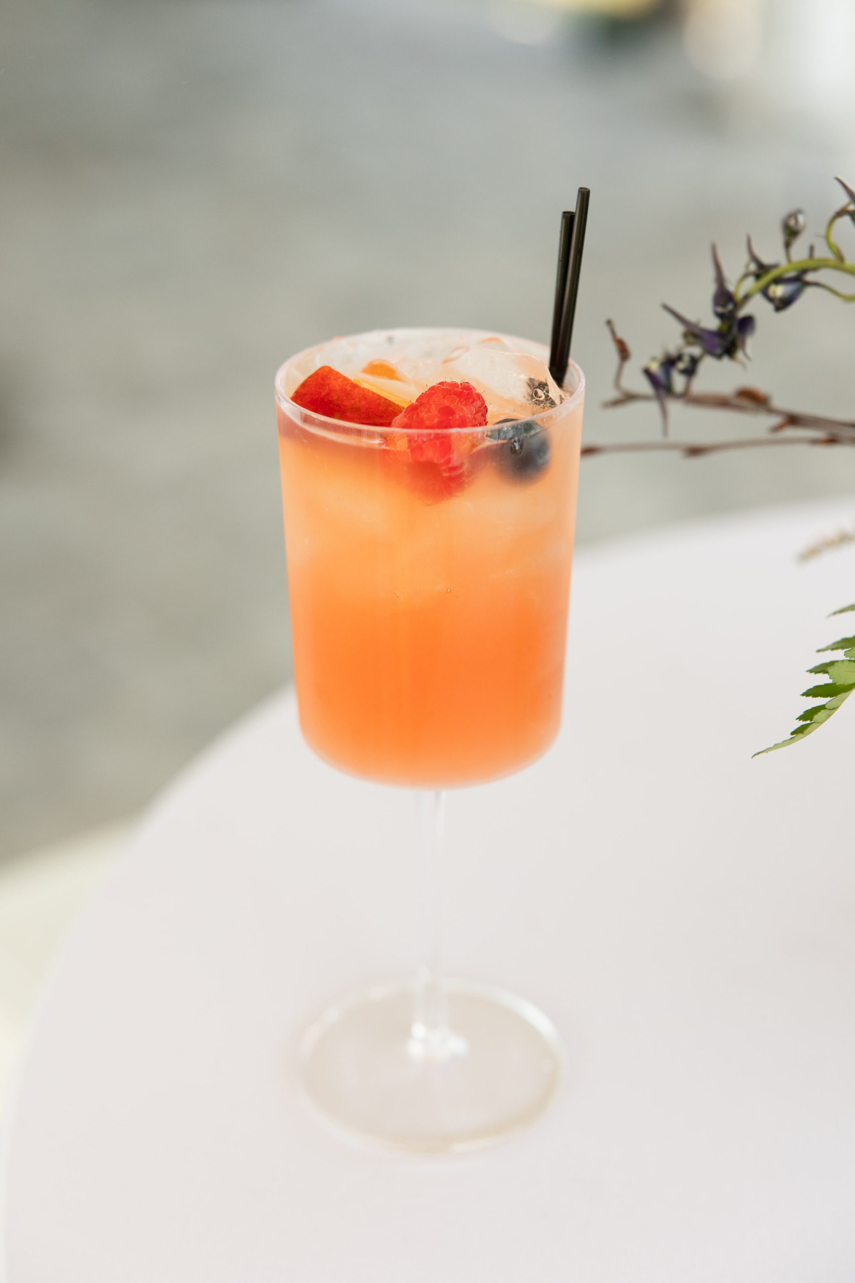 A tall glass of peach-colored cocktail garnished with ice cubes, strawberries, and a blueberry, with a black straw, placed on a white surface—perfect for the grand opening event.