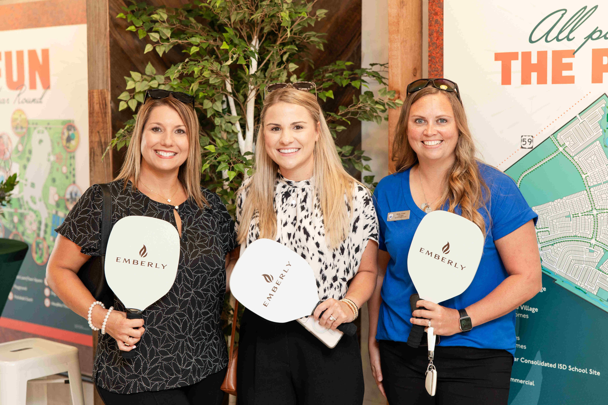 Three women standing together indoors, holding "Emberly" branded paddles and smiling, are in front of a display board with a map and text details at the grand opening of the information center.