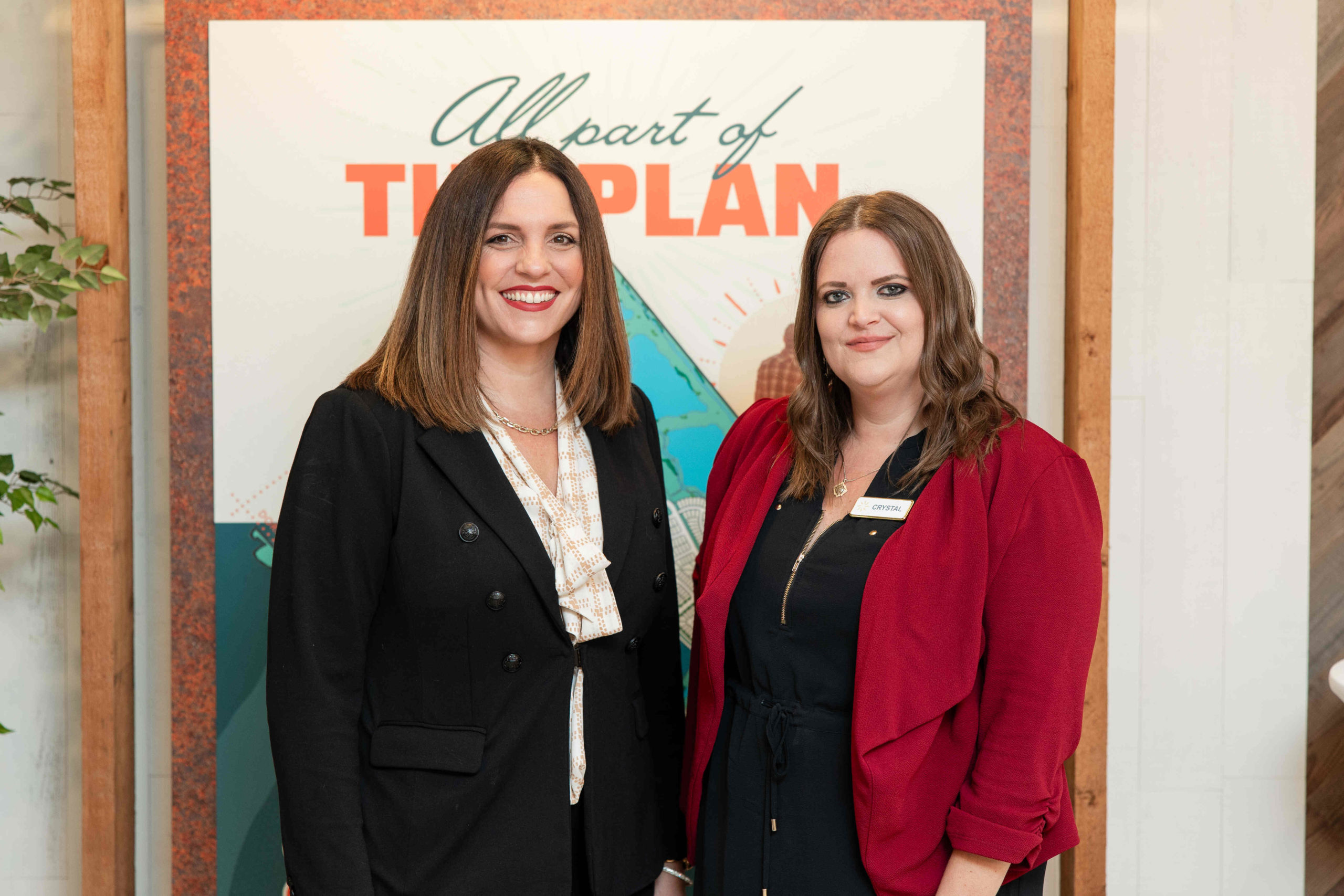 Two women stand smiling in front of a colorful poster that reads "All part of THE PLAN." One wears a black blazer and the other a red jacket. They are at the Grand Opening Event for the new Information Center.