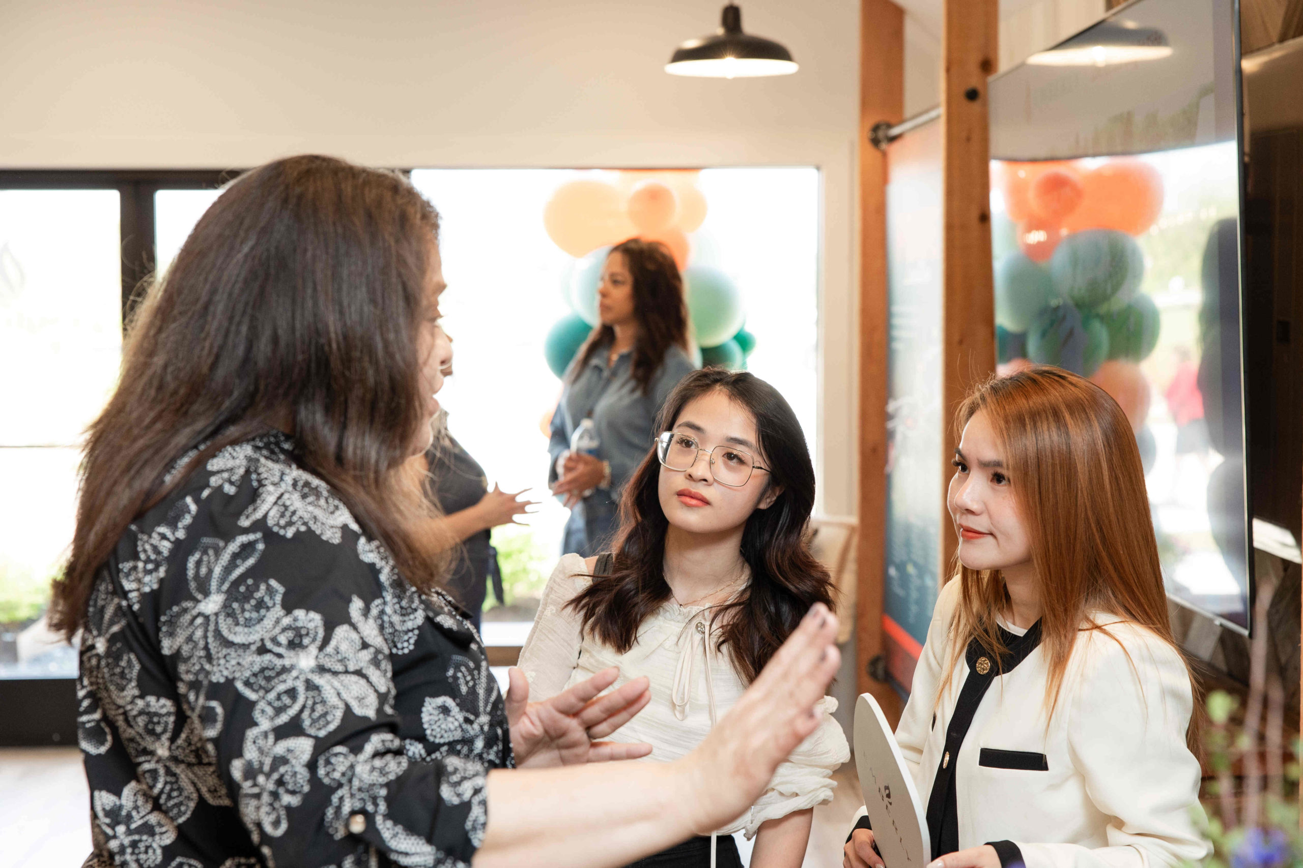 Three women are engaged in a conversation in a well-lit room with balloons in the background, likely attending an event. Two of them hold papers while listening attentively to the third woman.