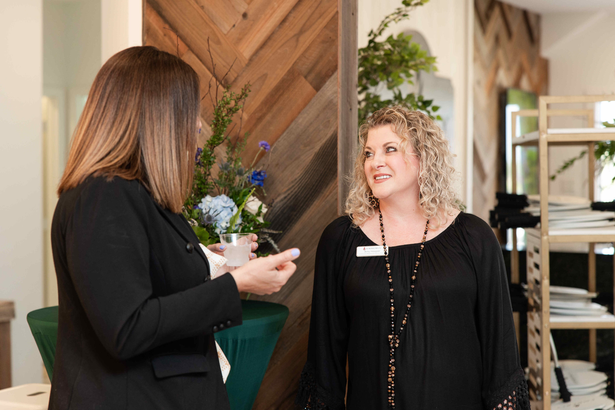 Two women are having a conversation in a room with wooden panels and green tablecloths, likely discussing the grand opening. One woman has straight brown hair and the other has curly blonde hair. Both are wearing black outfits, adding a touch of elegance to this event.