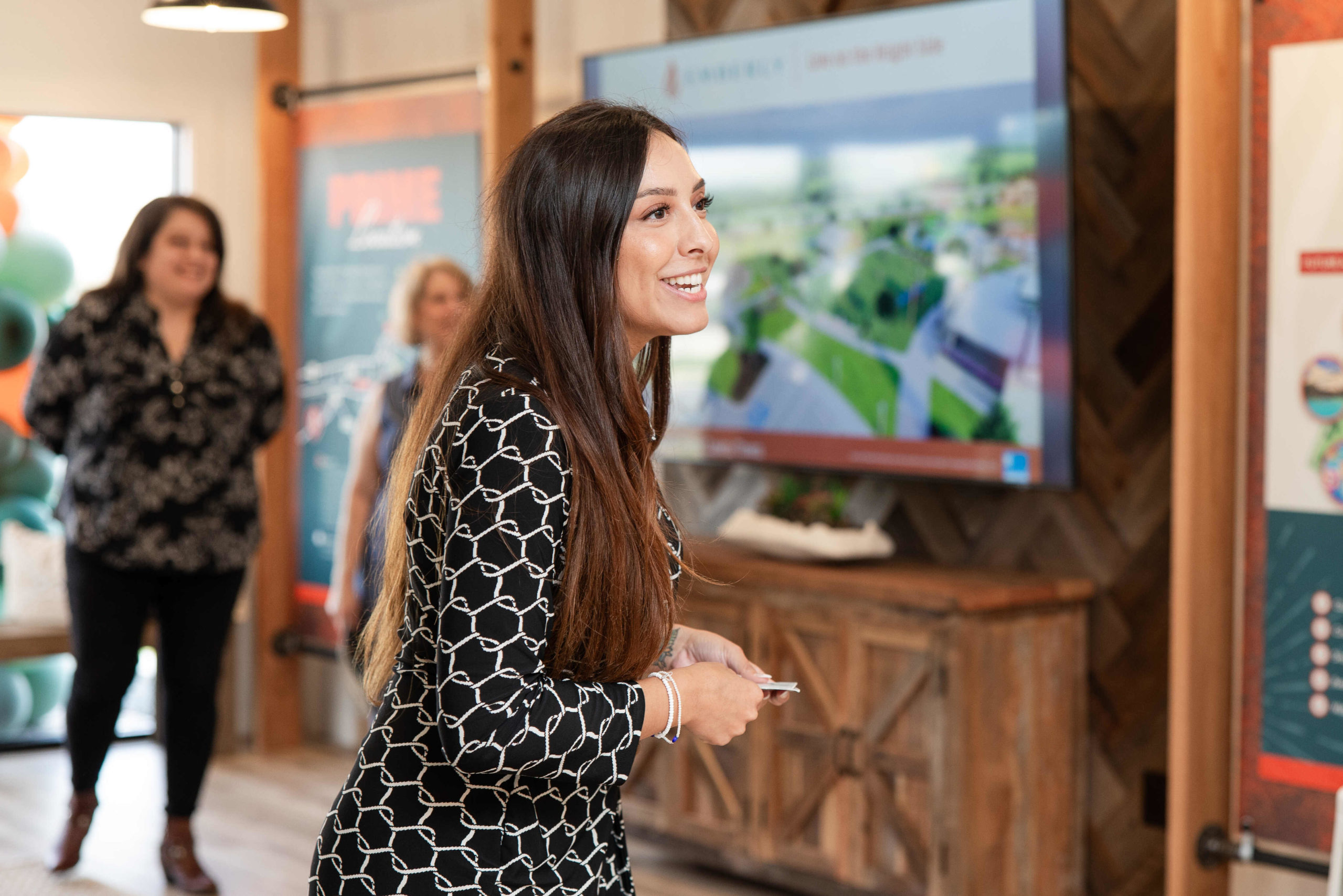 A woman dressed in black and white smiles while holding a small object in her hand at the Grand Opening Event, with two other people and a large screen displaying a presentation in the background.