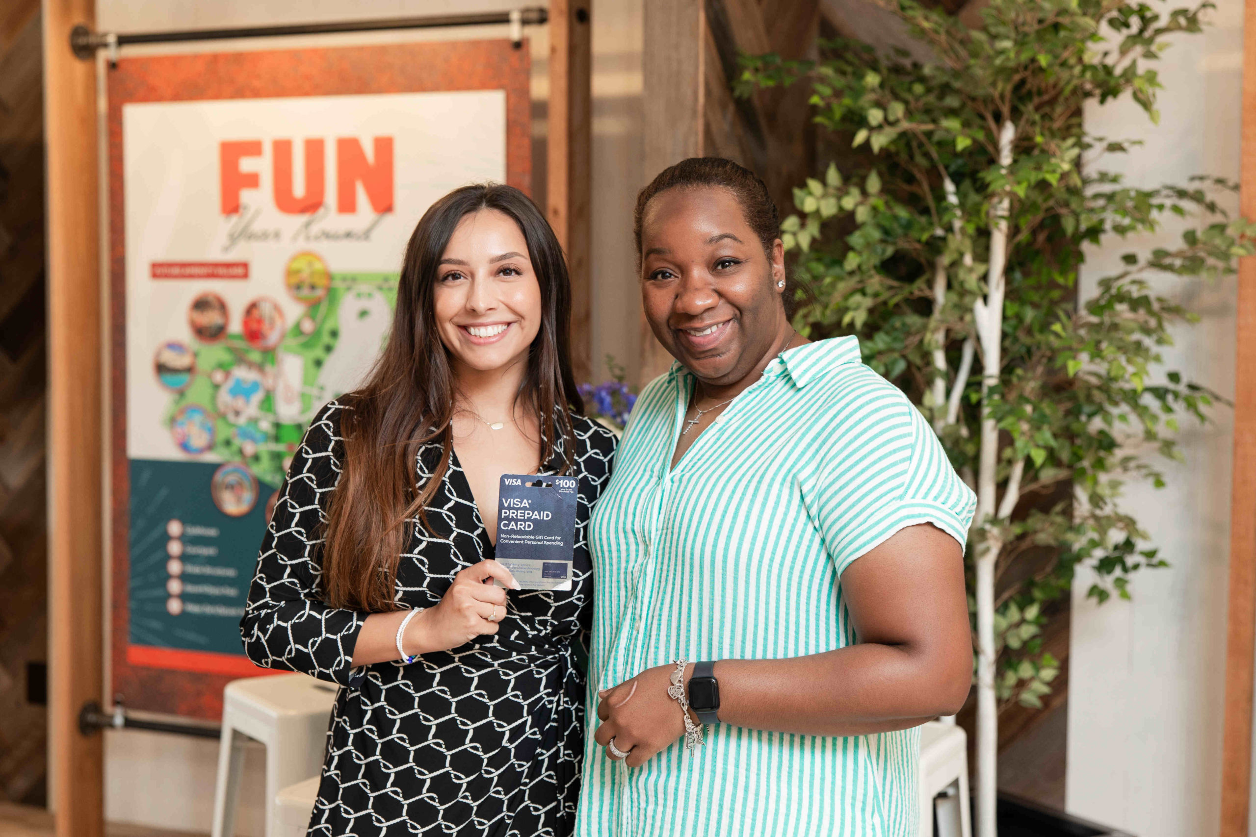 Two women smile at the camera, one holding a VISA gift card. A colorful poster with the word "FUN" is visible in the background, along with a tall plant, celebrating the event at the Information Center