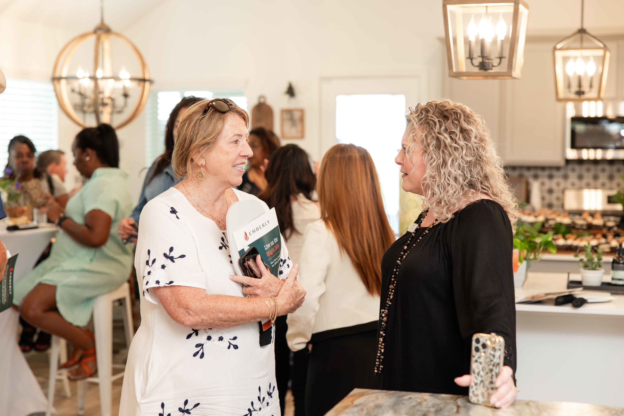 Two women are engaged in conversation at a social gathering in a brightly lit kitchen during the Grand Opening Event. Others mingle in the background, and various books and decorations are visible.