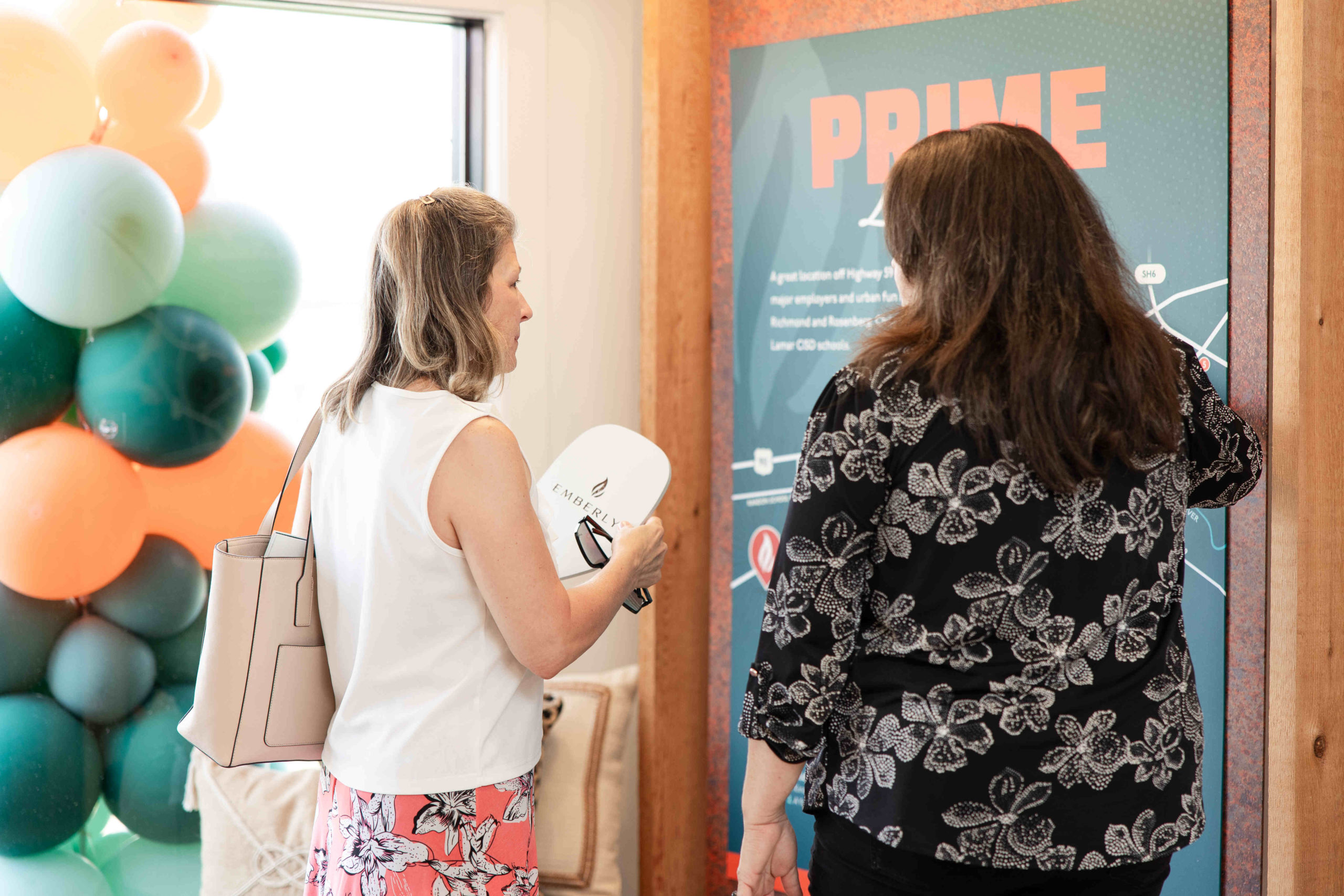 Two women examine a display board labeled "PRIME" at the Grand Opening Event. One points at the board while the other holds a white fan and a beige handbag. They stand next to a colorful balloon arrangement, highlighting the festive atmosphere of the Information Center