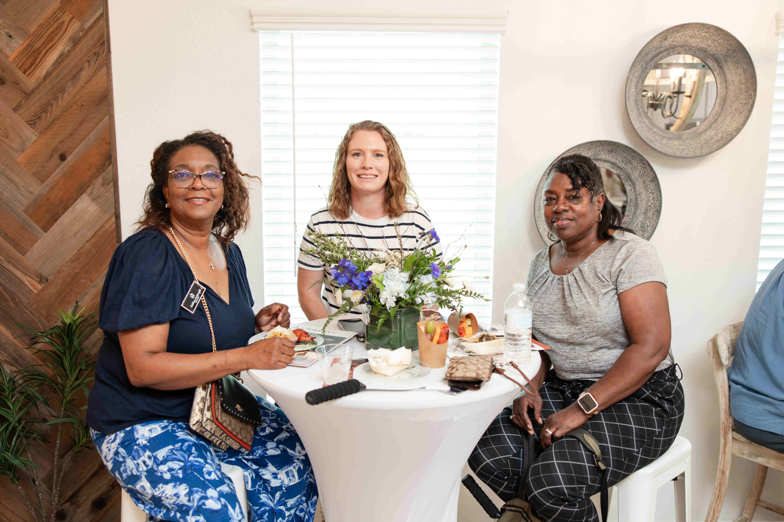 Three women seated around a tall table with food and drinks, smiling towards the camera. A floral centerpiece and a small bouquet are on the table, adding to the charm of this Grand Opening event.