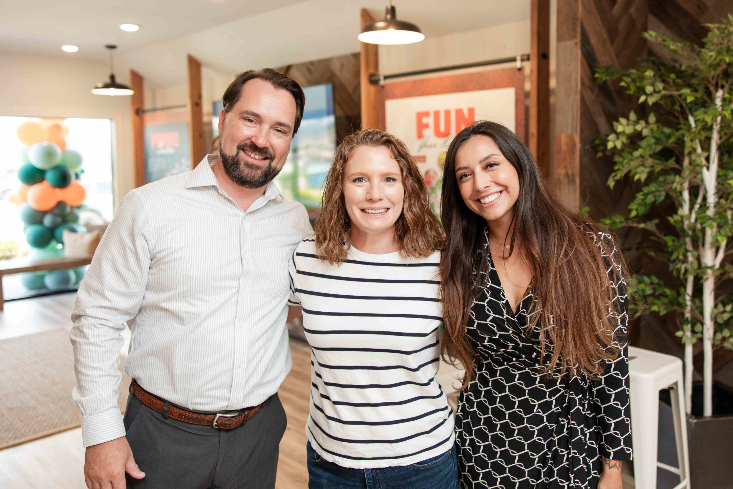 Three people stand indoors smiling at the camera; two women and one man. The space has wooden accents, visible greenery, and a sign that reads "FUN." It appears to be a festive event, possibly a grand opening of an information center.