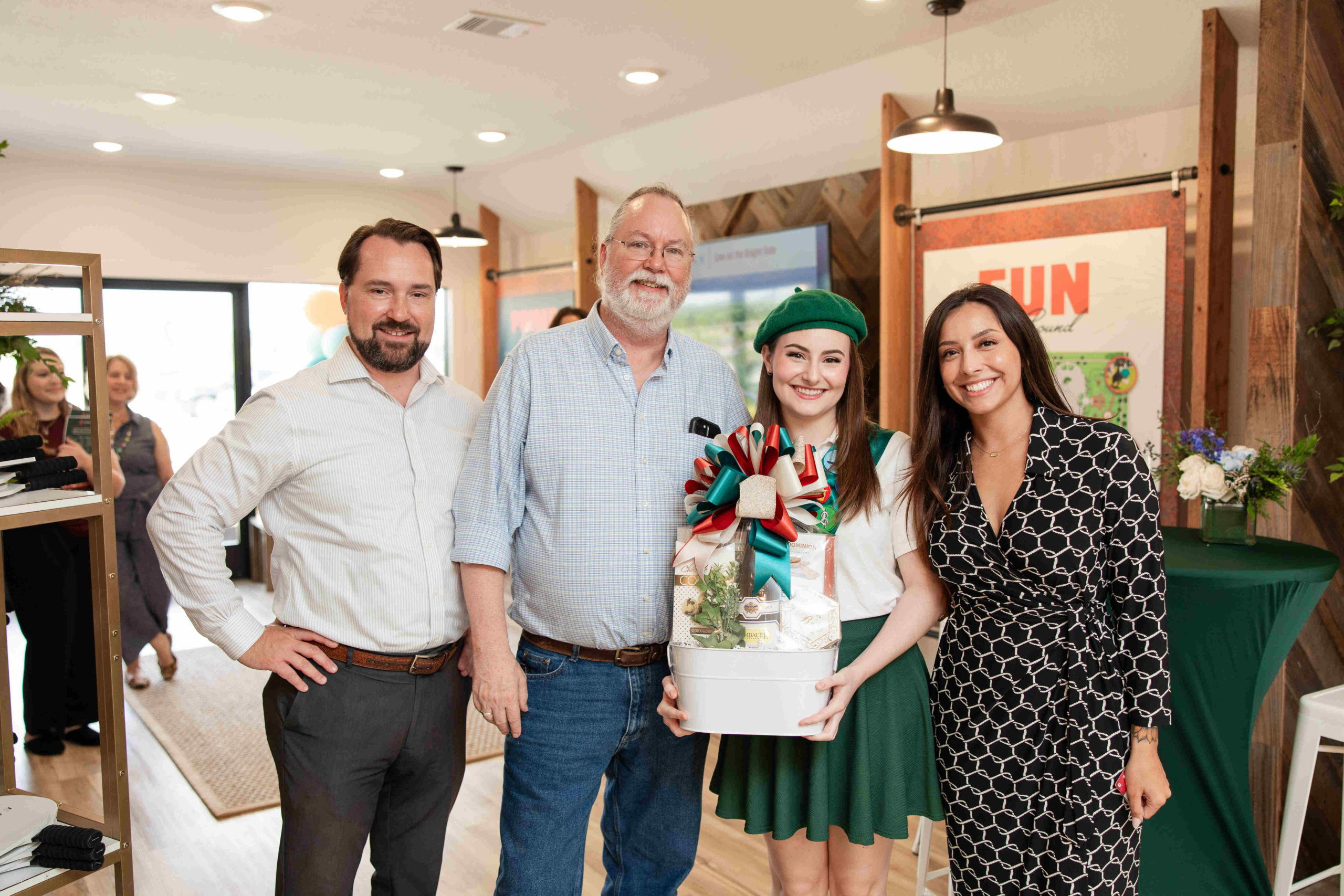 Four people standing indoors at an event, smiling at the camera; one holds a gift basket adorned with ribbons, celebrating the grand opening.