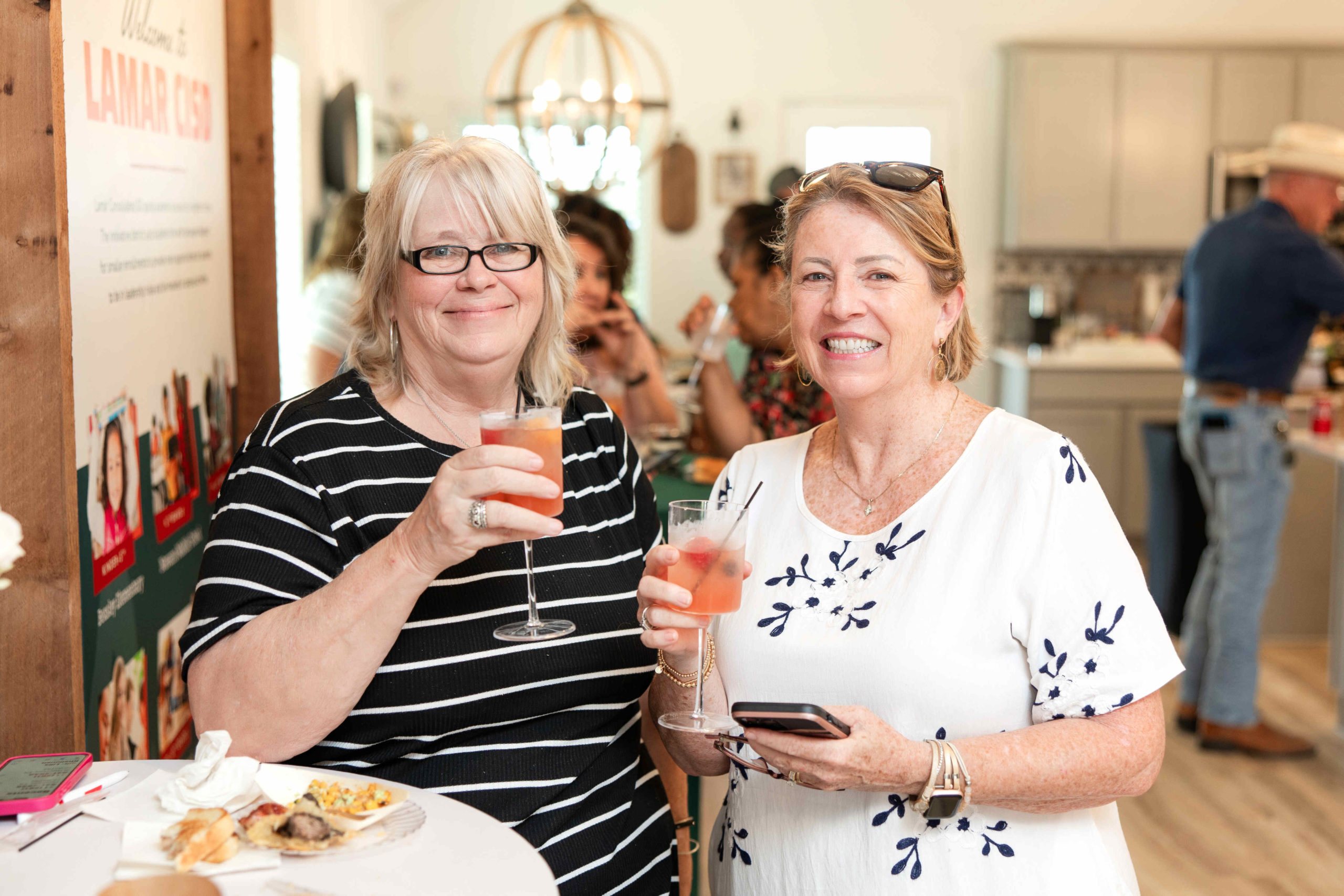 Two women standing at a table holding drinks and smiling at the grand opening event in a well-lit room with other people and food in the background.