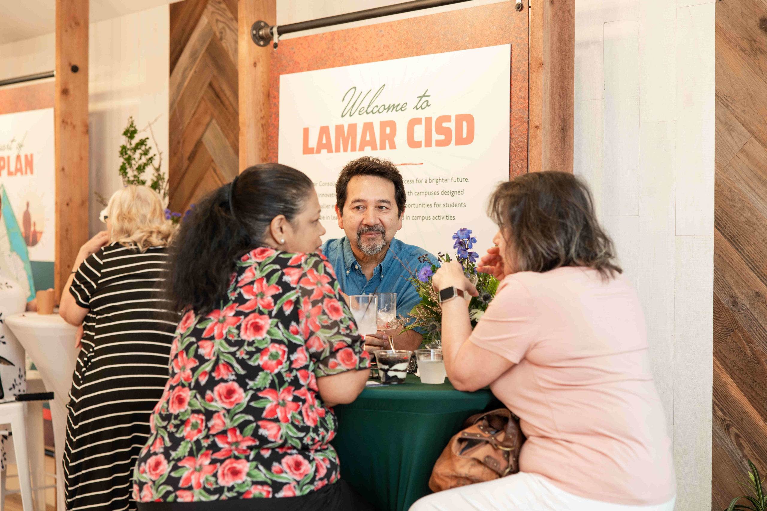 Three people converse at a table in front of a "Welcome to Lamar CISD" sign, with another person in the background, setting the stage for the Grand Opening of the Information Center.