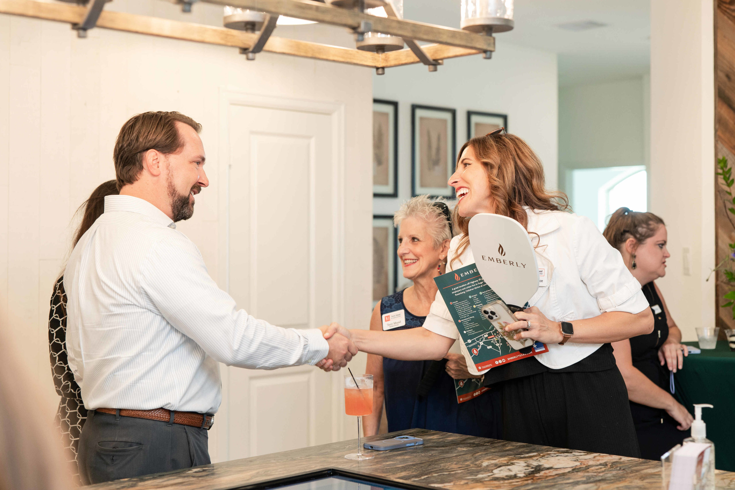 Two people engage in a handshake at an indoor gathering. Another person in the background holds a brochure and paddle, likely for the event