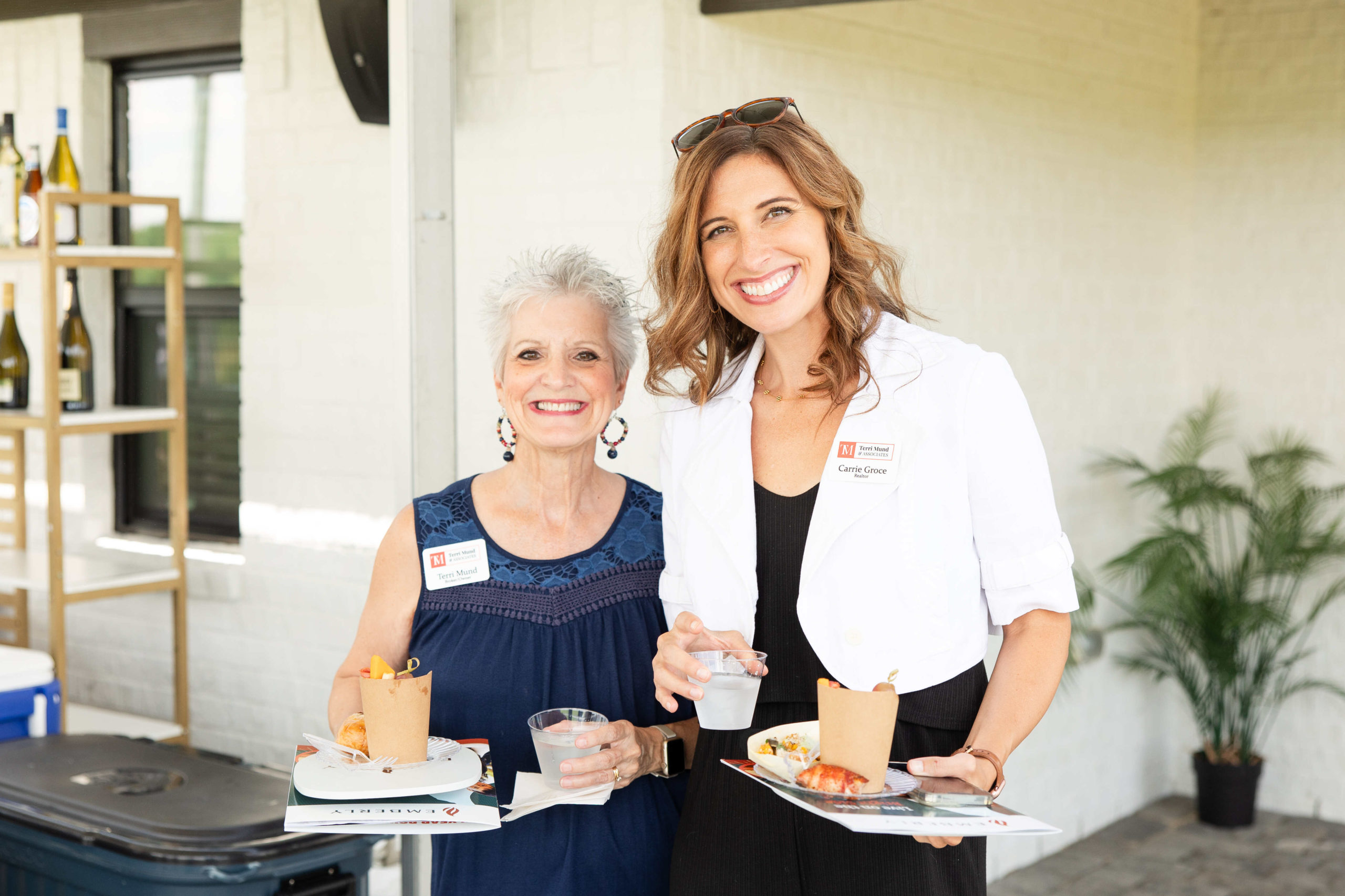 Two women smiling, holding plates of food and beverages at an outdoor event. One wears a white blazer with a name tag, hinting she might be from the Information Center, while the other wears a dark blue sleeveless top. It