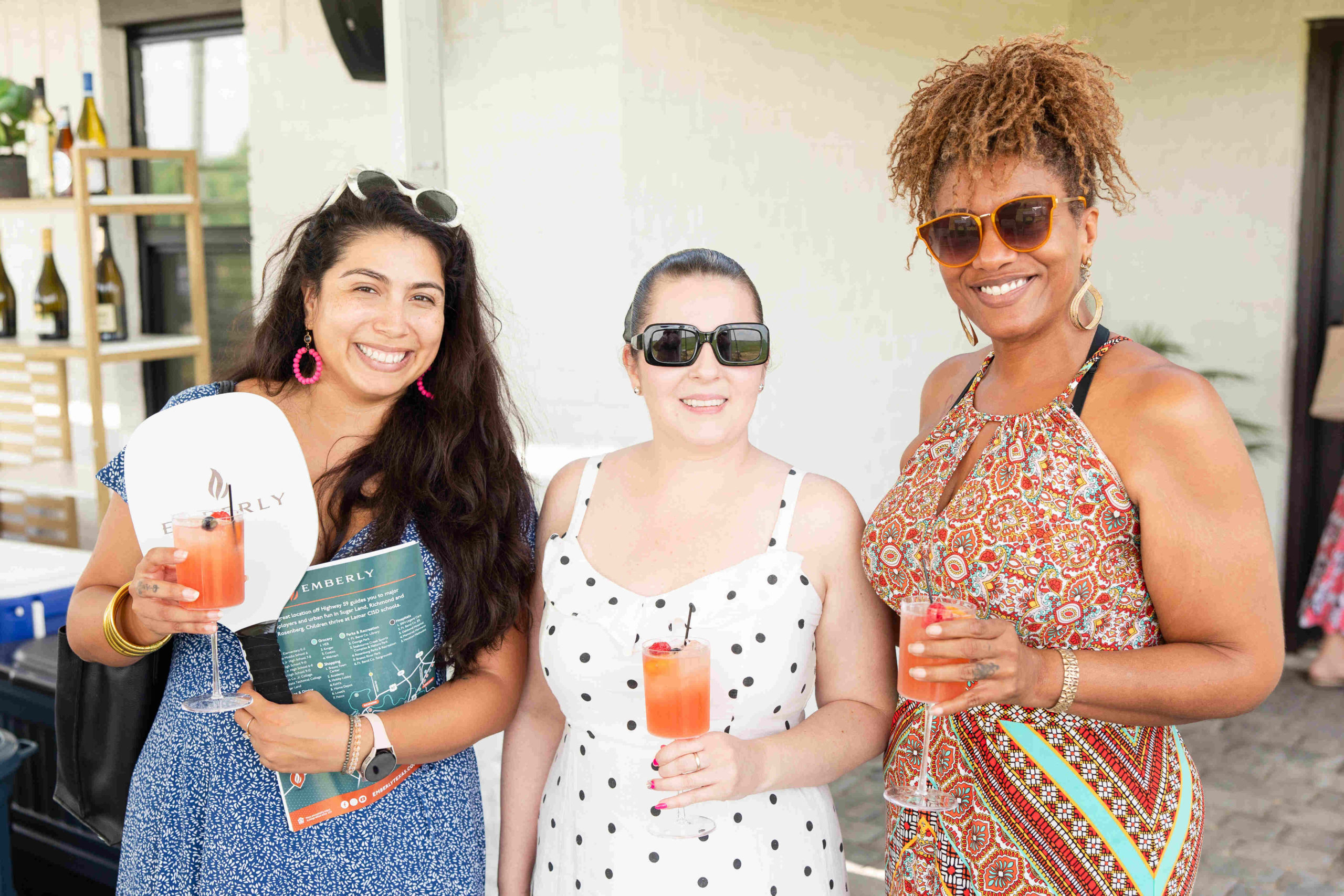 Three women smiling at the camera while holding drinks at the event. They are dressed in summer attire, with one wearing sunglasses and holding a booklet about the Grand Opening.