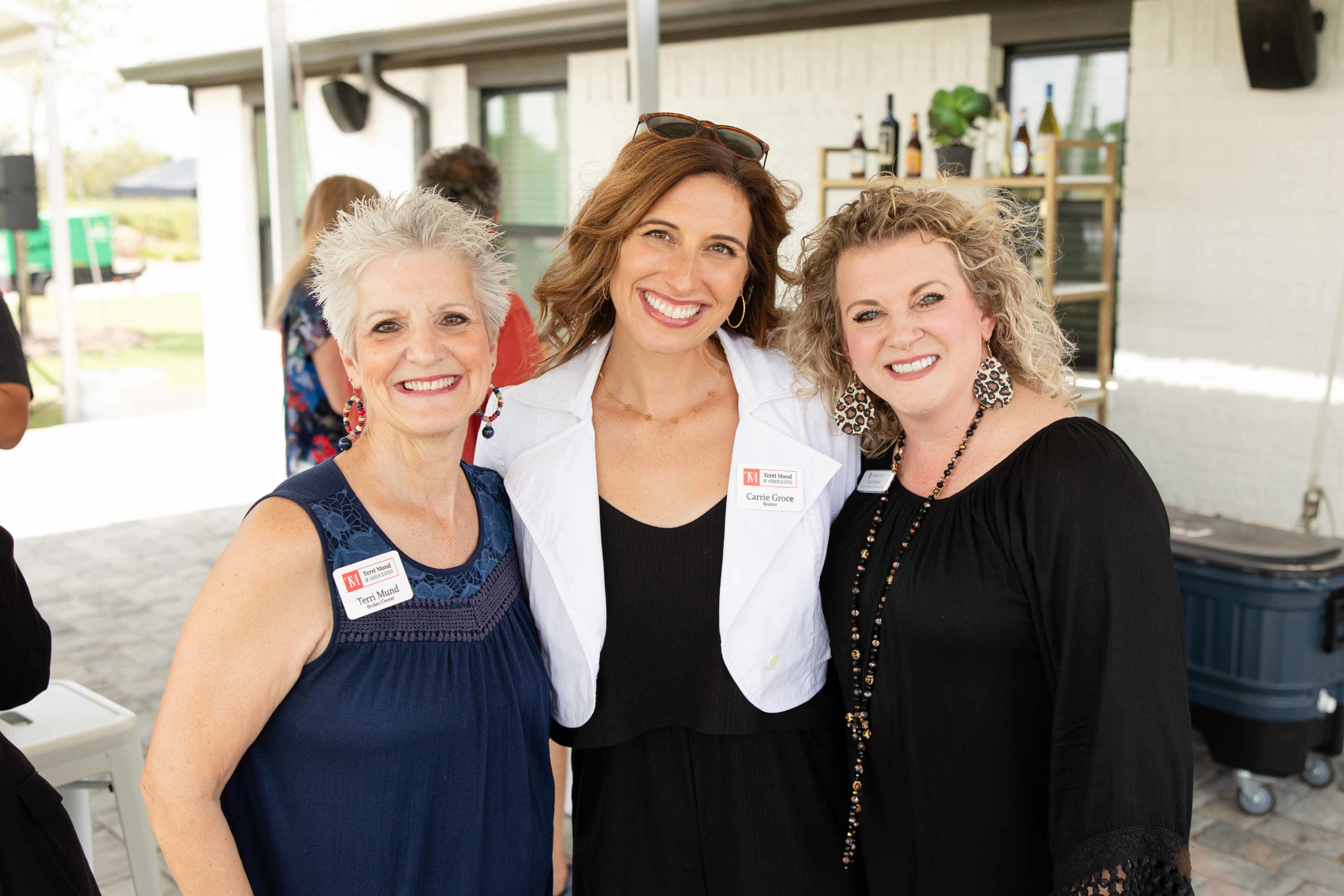 Three women are standing and smiling at an outdoor event. They are wearing name tags and are dressed in casual attire. A building and some people are visible in the background, possibly indicating the grand opening of a new Information Center.