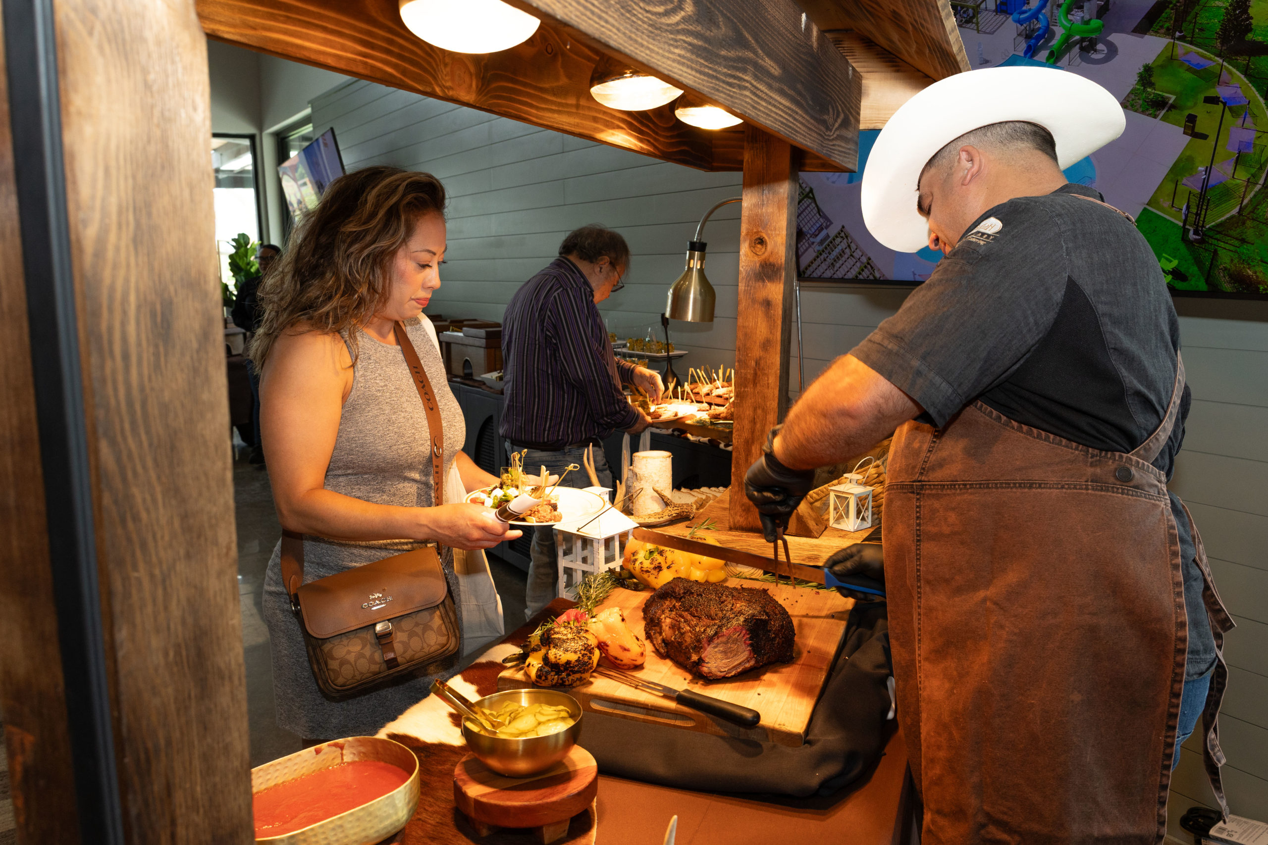 At the Grand Opening of Haven Amenity Village, a man in an apron and cowboy hat slices meat at a carving station while a woman serves herself food from a buffet line.