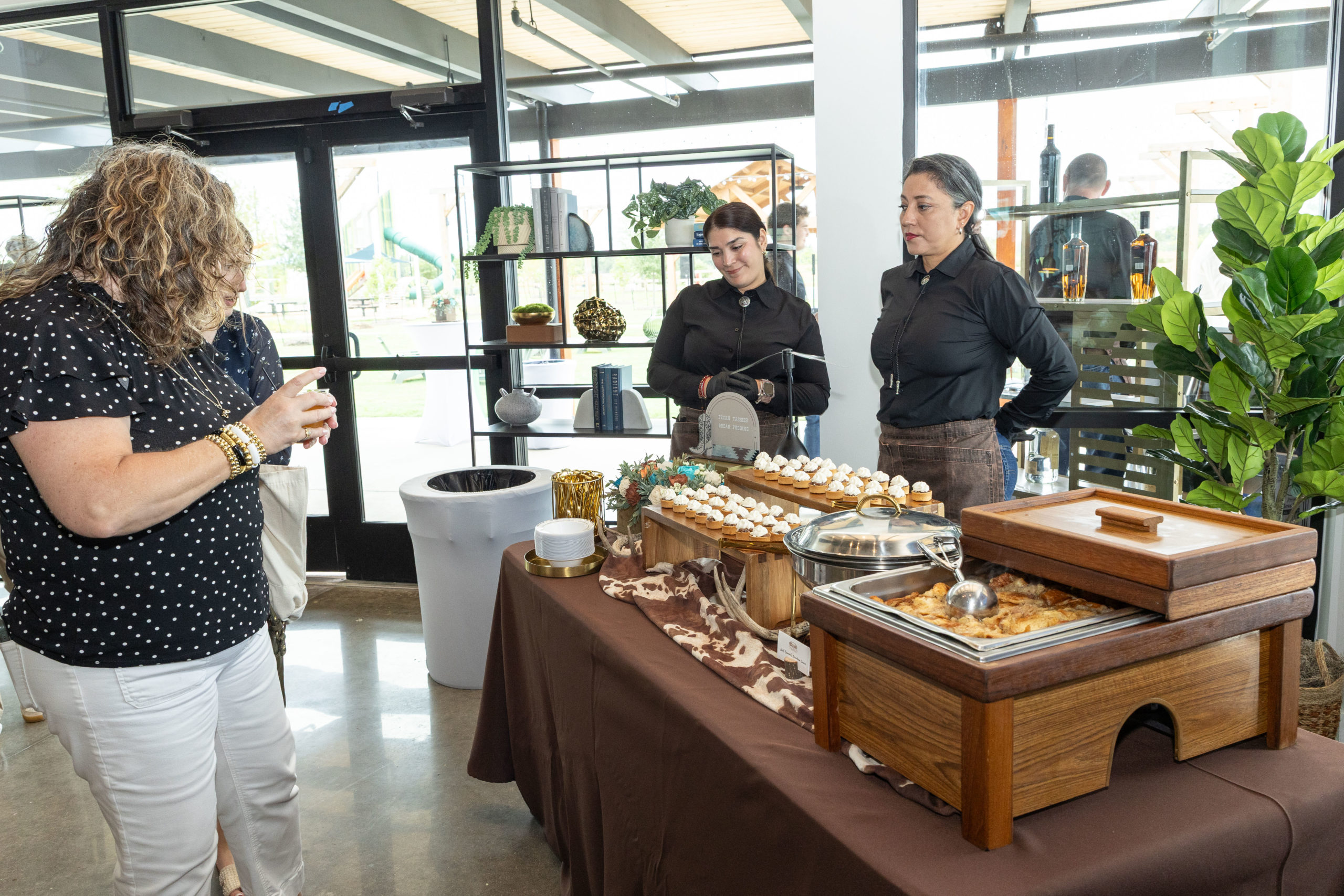 At the Grand Opening of Haven Amenity Village, a woman stands in front of a buffet table with food and desserts, attended by two servers dressed in black uniforms in a bright indoor space.