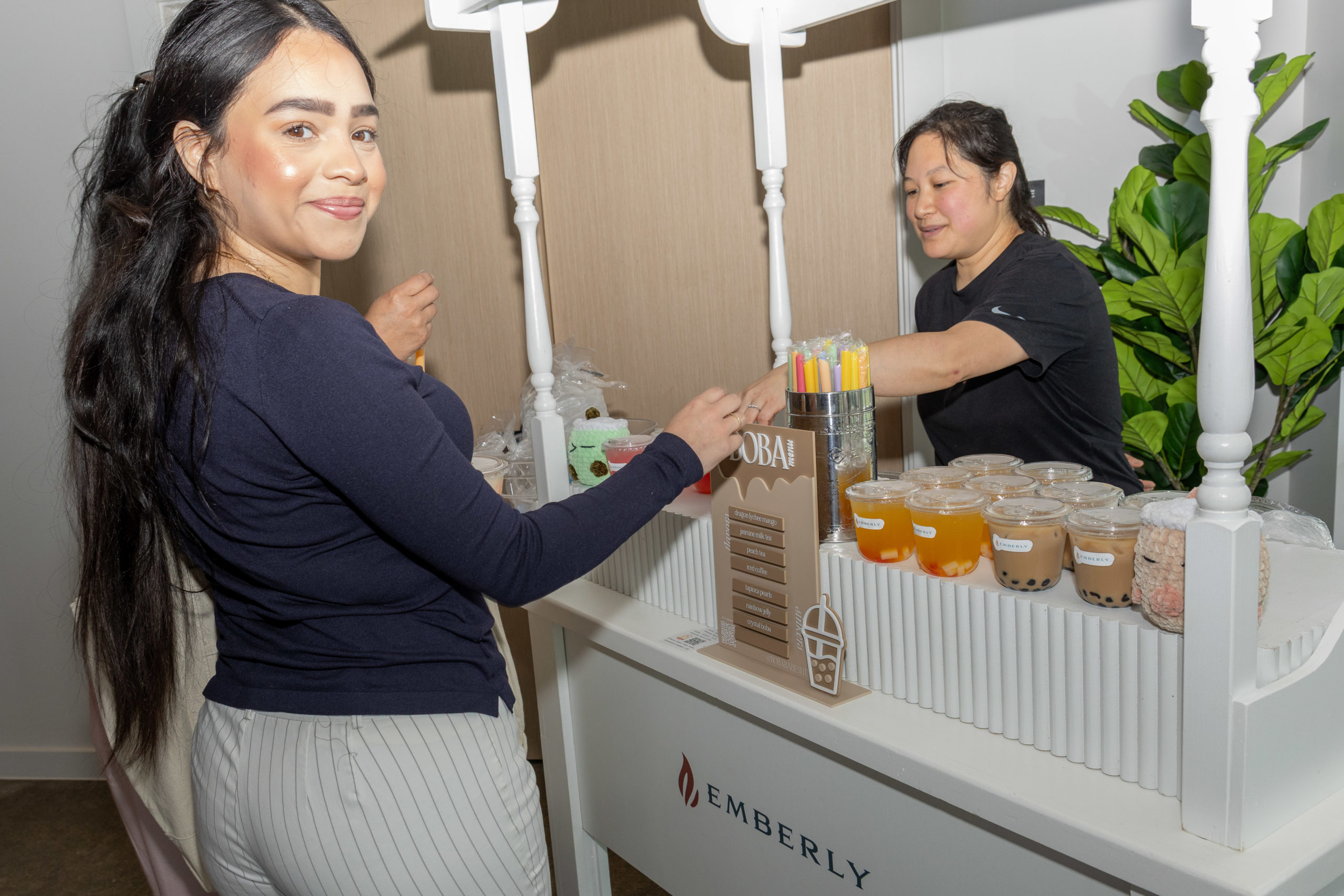 At the Haven Amenity Village Grand Opening, two women stand at a bubble tea cart; one serves drinks while the other selects a cup, surrounded by bubble tea cups and colorful straws on the counter.