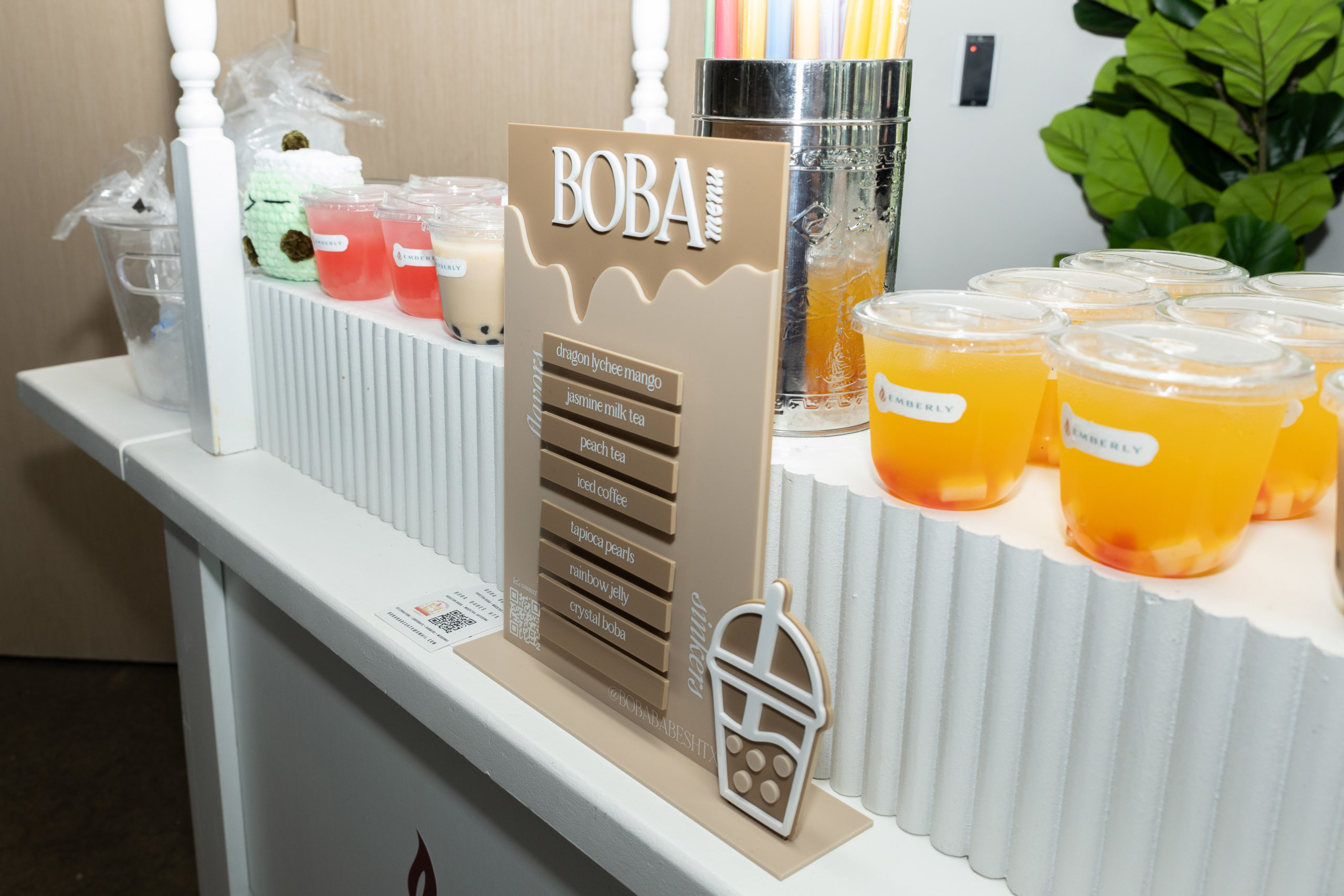 At the Haven Amenity Village Grand Opening, a boba tea stand displays a menu and several cups of boba tea with fruit pieces, alongside a container of colorful straws and a leafy plant in the background.