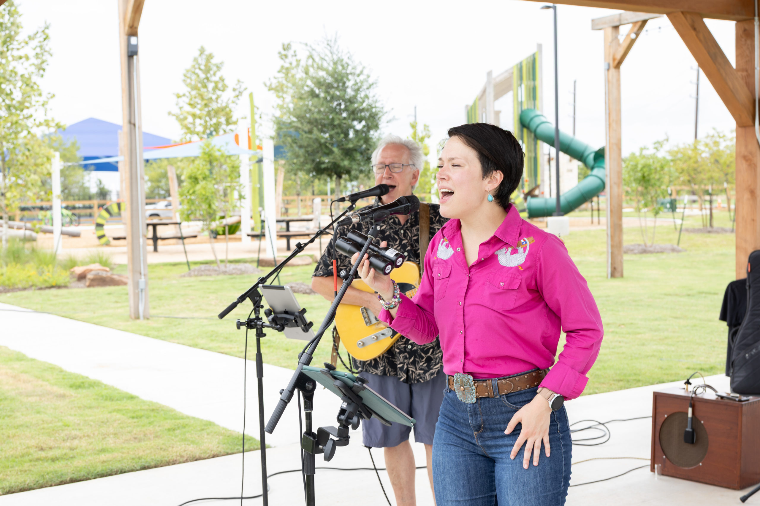 A woman sings into a microphone while a man plays guitar beside her at the Grand Opening of Haven Amenity Village, held at an outdoor covered pavilion with playground equipment in the background.
