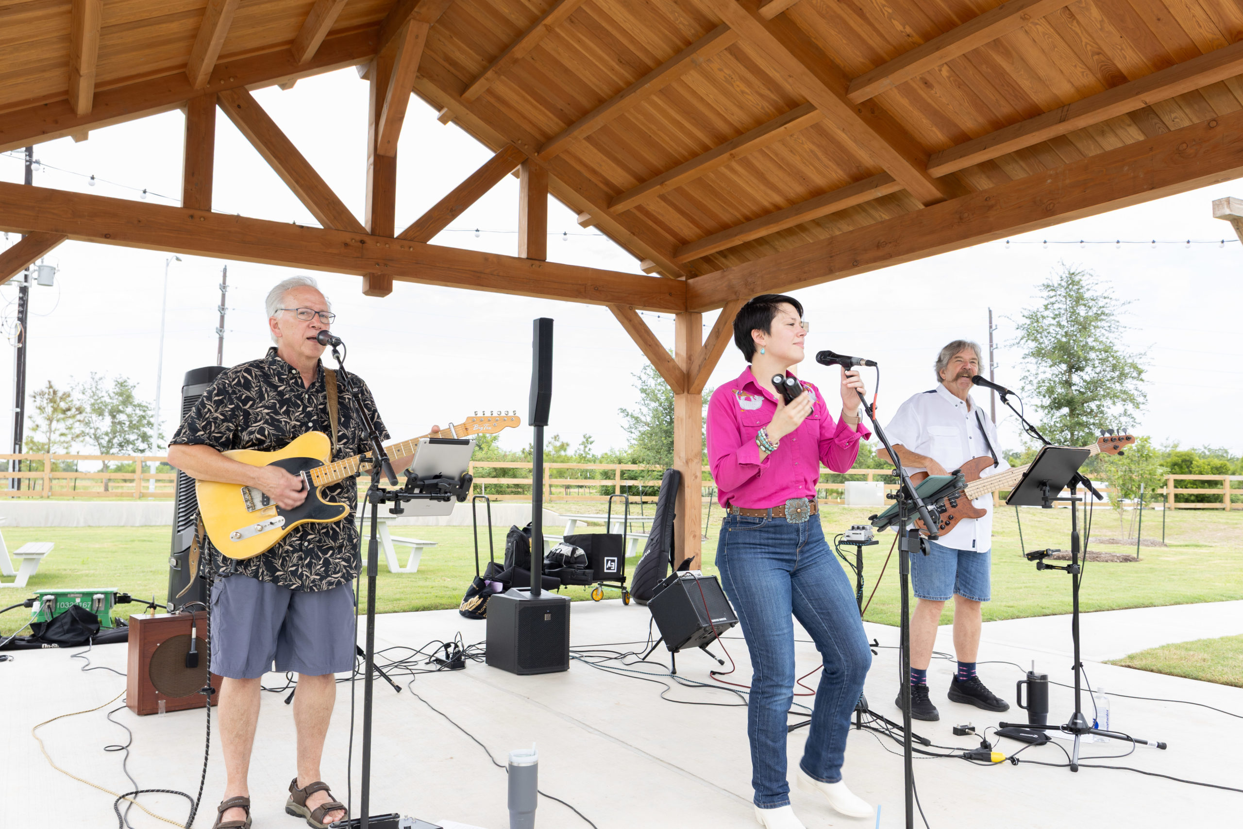 Three musicians perform under a wooden pavilion at the Haven Amenity Village Grand Opening; one plays guitar and sings, another sings into a microphone, and the third plays bass guitar. The outdoor setting features green grass and trees.