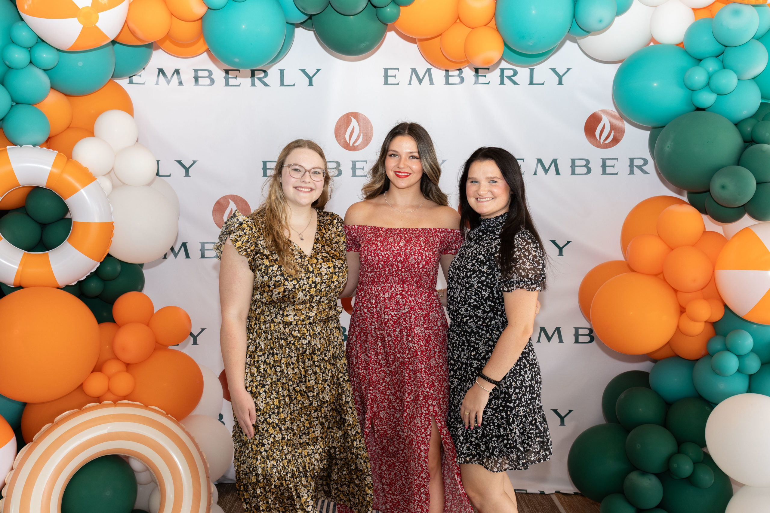 Three women stand in front of a backdrop with the word "EMBERLY," surrounded by orange, green, and white balloons, celebrating the Grand Opening of Haven Amenity Village.